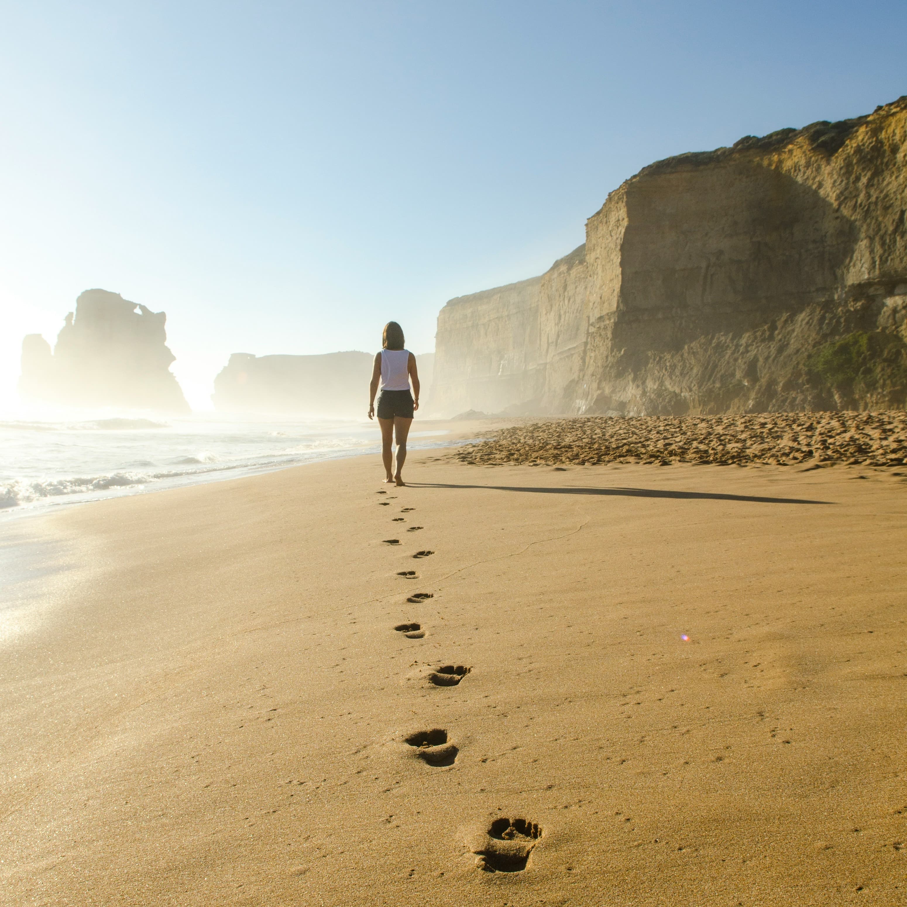 A photo of a woman walking on the beach. She is walking close to the water, away from the camera and ahead of her are some cliffs. Behind her, she is leaving footprints.
