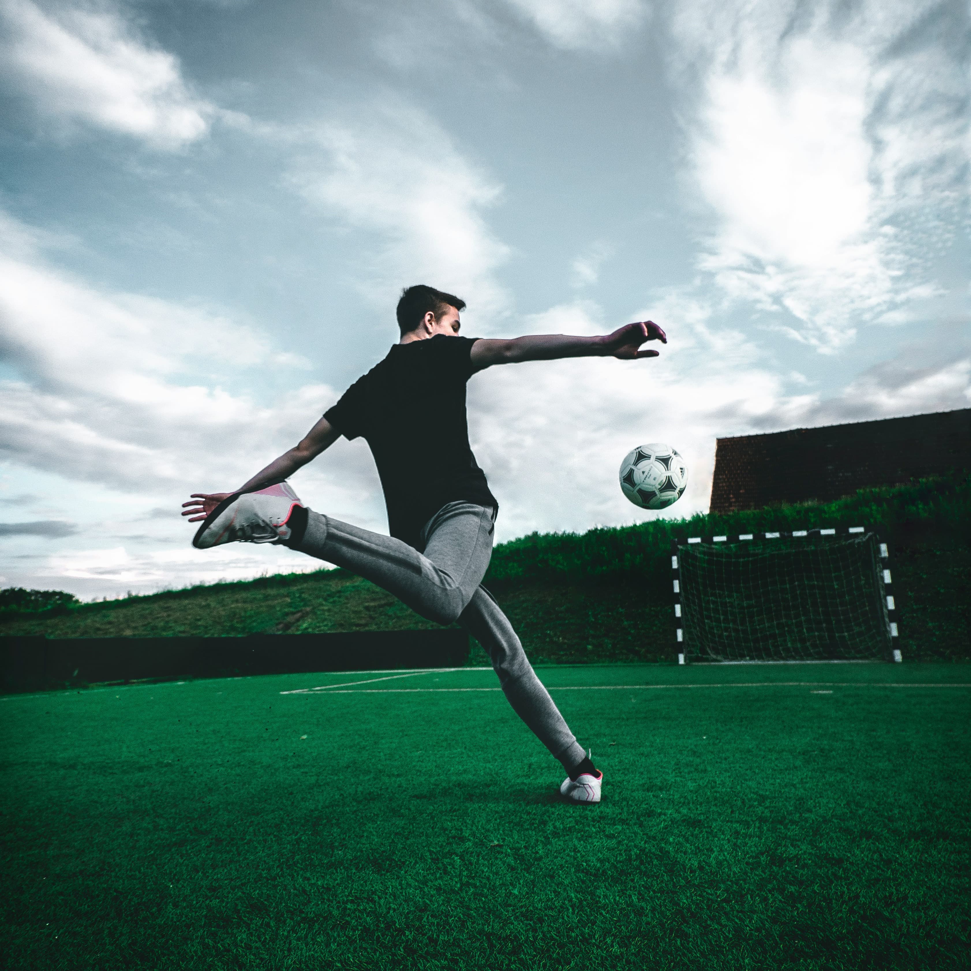 A dramatic photo of a soccer player, taken from behind. The ball is in midair and he right leg is preparing to kick it in the direction of a goal.