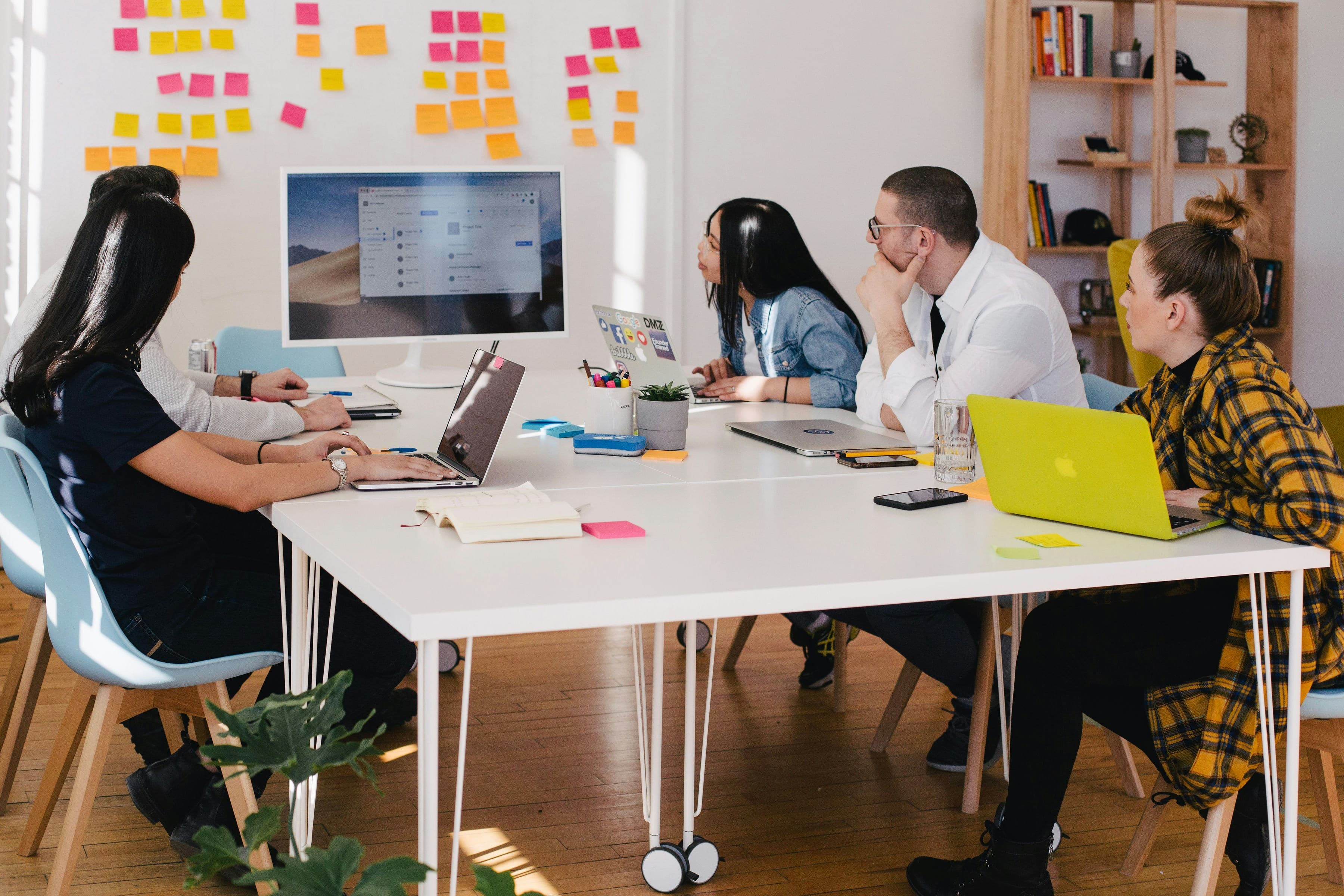 A photo of a group of people gathered around a white conference table. They are looking towards the far end of the table where there is a computer monitor and, on the wall behind it, several post-it notes in different colors. The people at the table, from right-to-left: A woman in a plaid yellow jacket and black pants with brown hair in a messy bun. On the table, she has a Macbook with a lemon yellow skin, a glass of water, and a black smartphone. Next, a man with a buzz cut, dark hair, and glasses wearing a white collared shirt over a black t-shirt. He has a closed silver Macbook and a smartphone on the table and is holding his chin in his hand. Next, a woman with long black hair and red lipstick is leaning forward. She's wearing a denim shirt and typing on a silver Macbook with several stickers on it. On the left side of the table, we can't se much, besides that a woman with long hair in dark clothes is sitting closer to us, blocking our view of a man in a light-colored neutral sweater. The woman is typing on an open laptop and has a notebook open beside her.