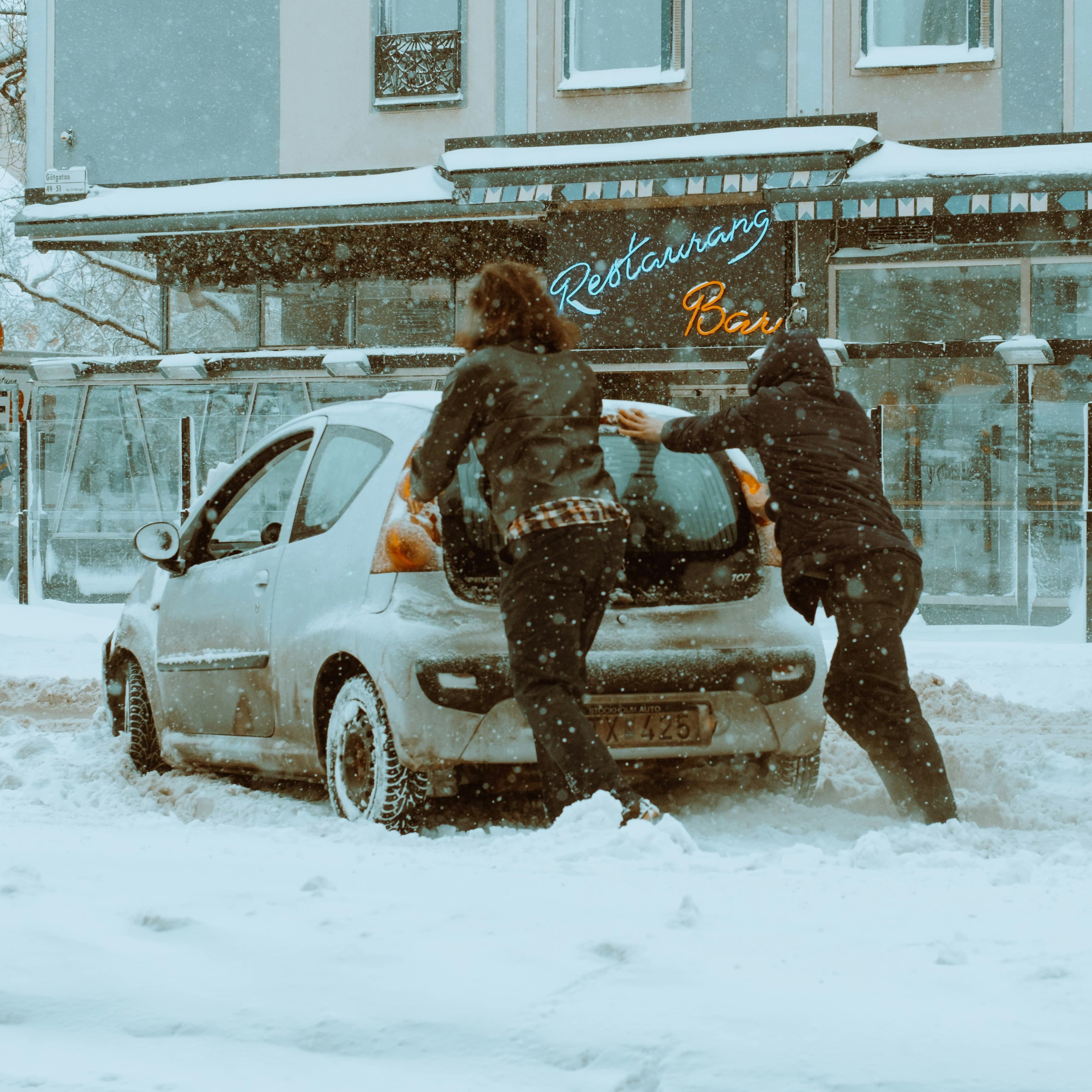 A photo of two people pushing a small dirty car down a street in a snowstorm.