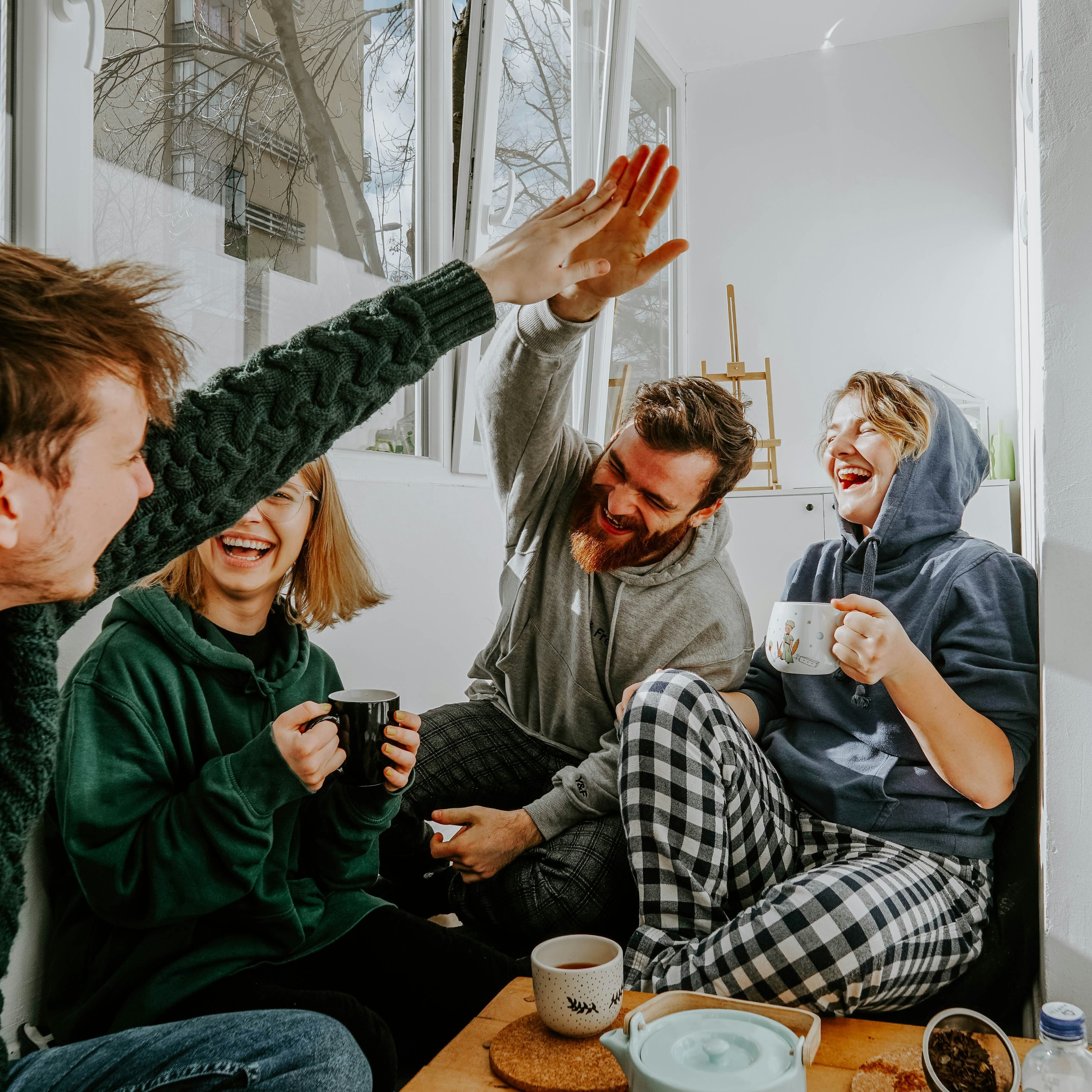 A group of three young white people are sitting around a coffee table and laughing in a largely white apartment. The two men are high-fiving across the table while the two women laugh, holding mugs. On the coffee table we can see a teapot and the men's mugs. All four are wearing loungewear. The window to the apartment is cracked open and beyond it we can see a winter tree and a high-rise apartment building.
