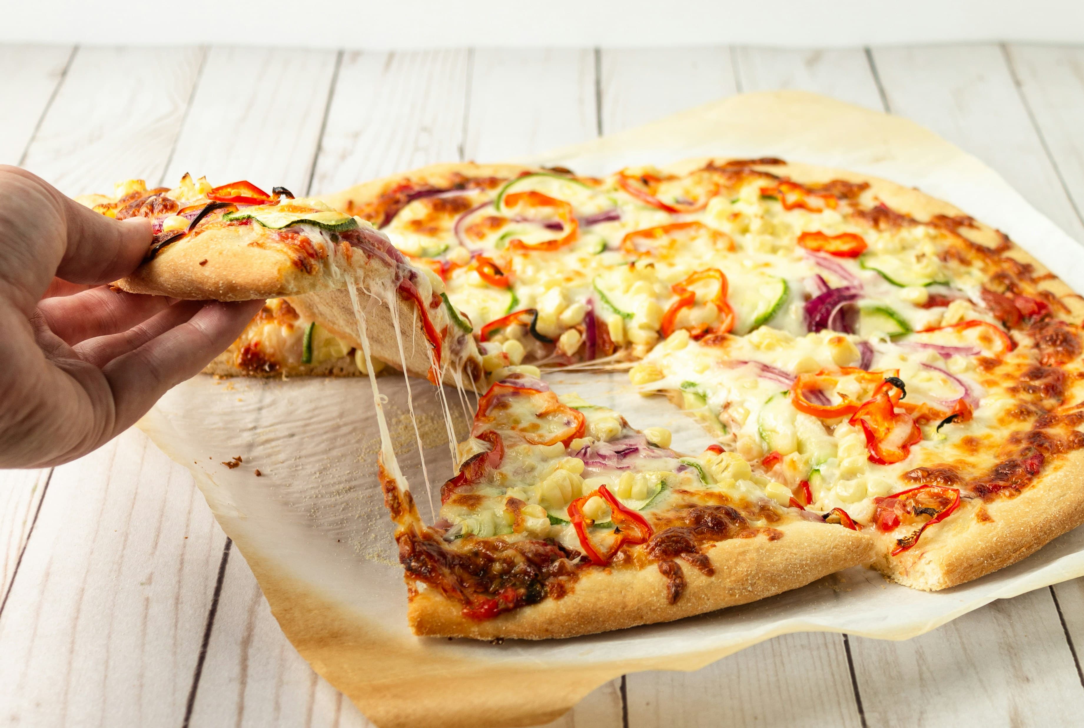 A photo of a vegetarian pizza on a sheet of parchment paper. A hand is reaching down to pick up one slice.