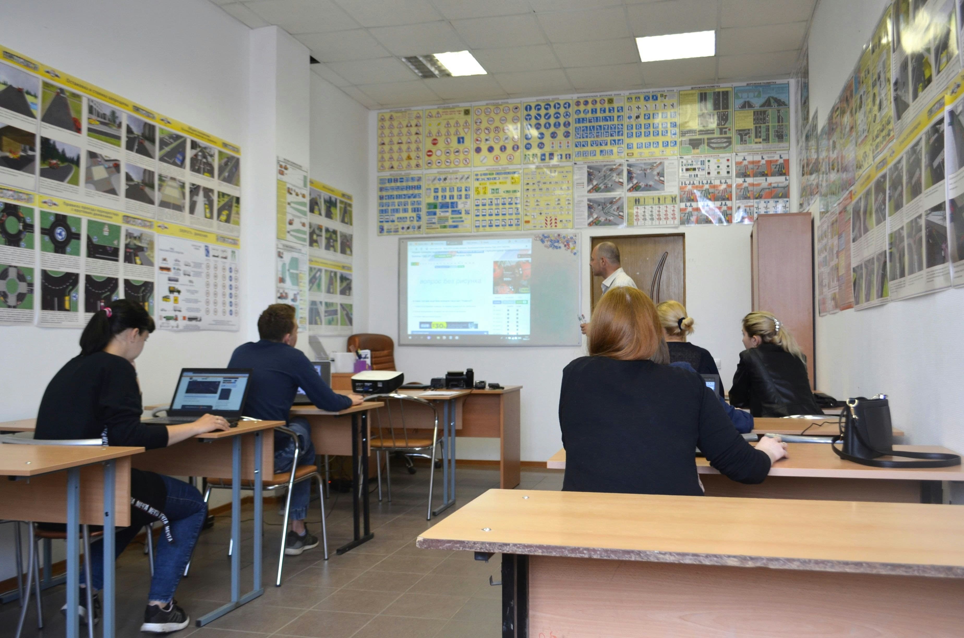 A small group of adult students sits at desks in a classroom. The walls are covered in educational diagrams and at the front of the room a teacher speaks in front of a small projector screen.