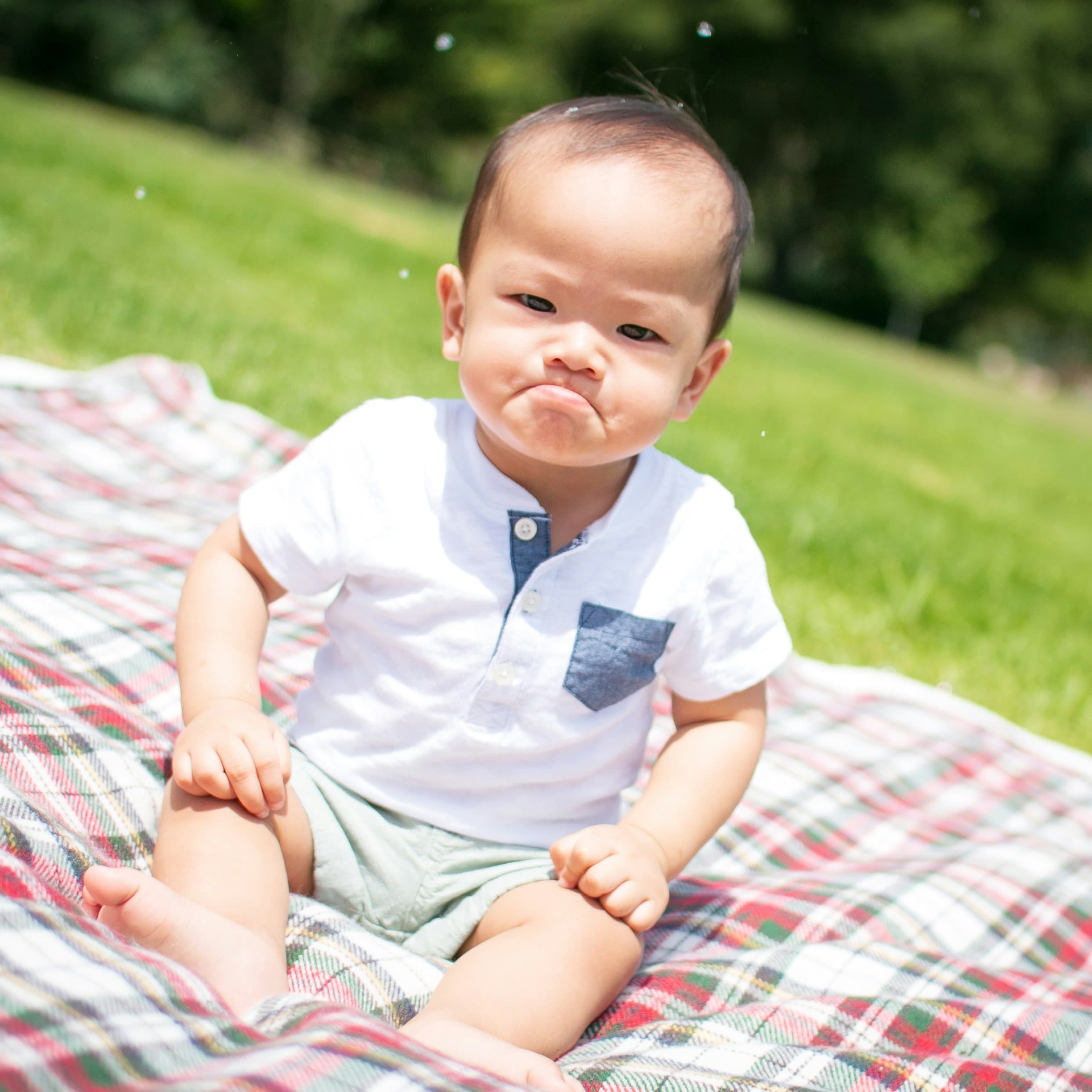 A baby with dark hair sits on a picnic blanket in a park. He has a comically huge frown on his face as he looks at the camera.
