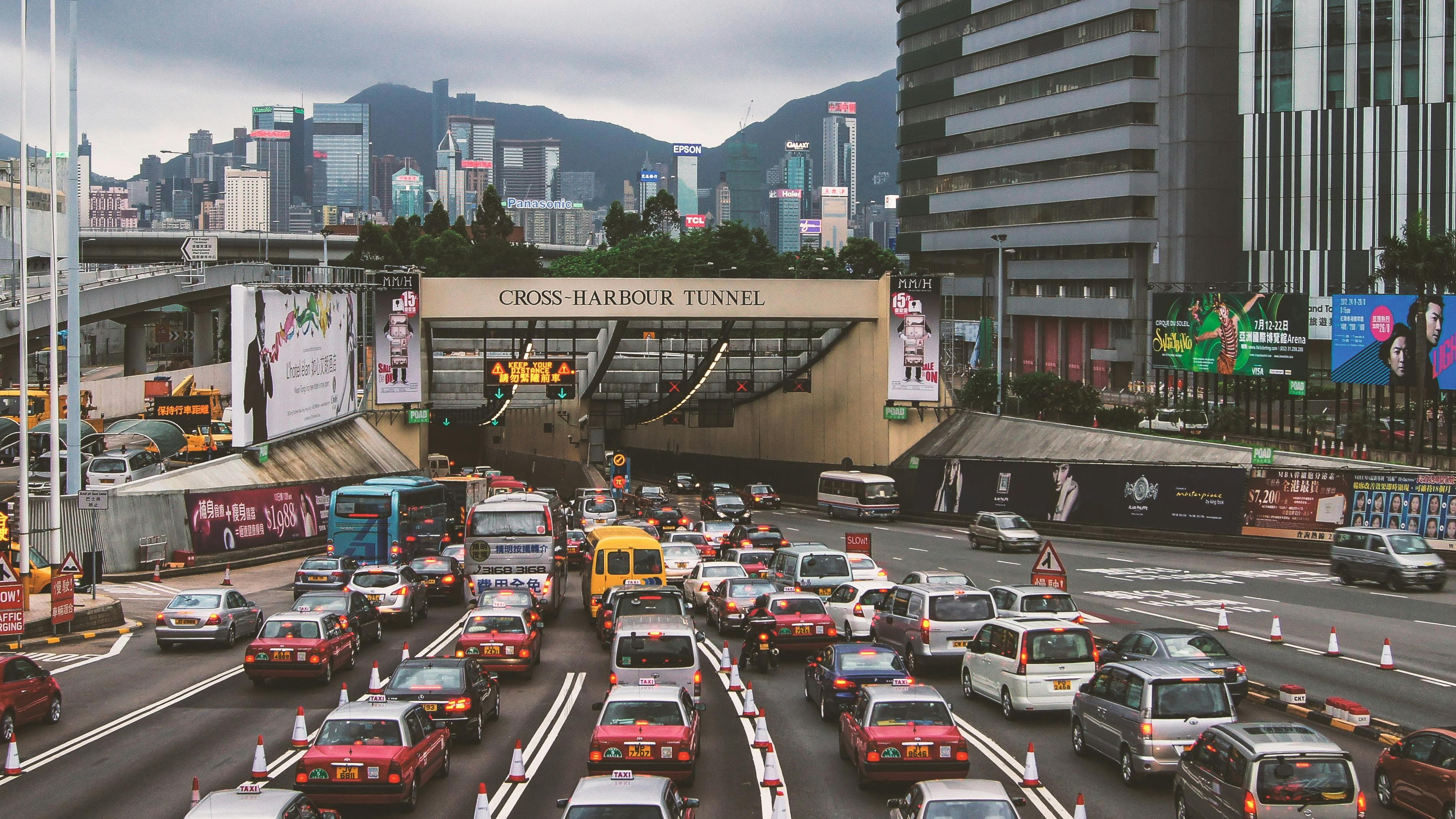 A photo of bumper-to-bumper traffic on a multi-lane highway. People are heading towards the Cross-Harbour Tunnel and an urban skyline can be seen in the background in font of some mountains.