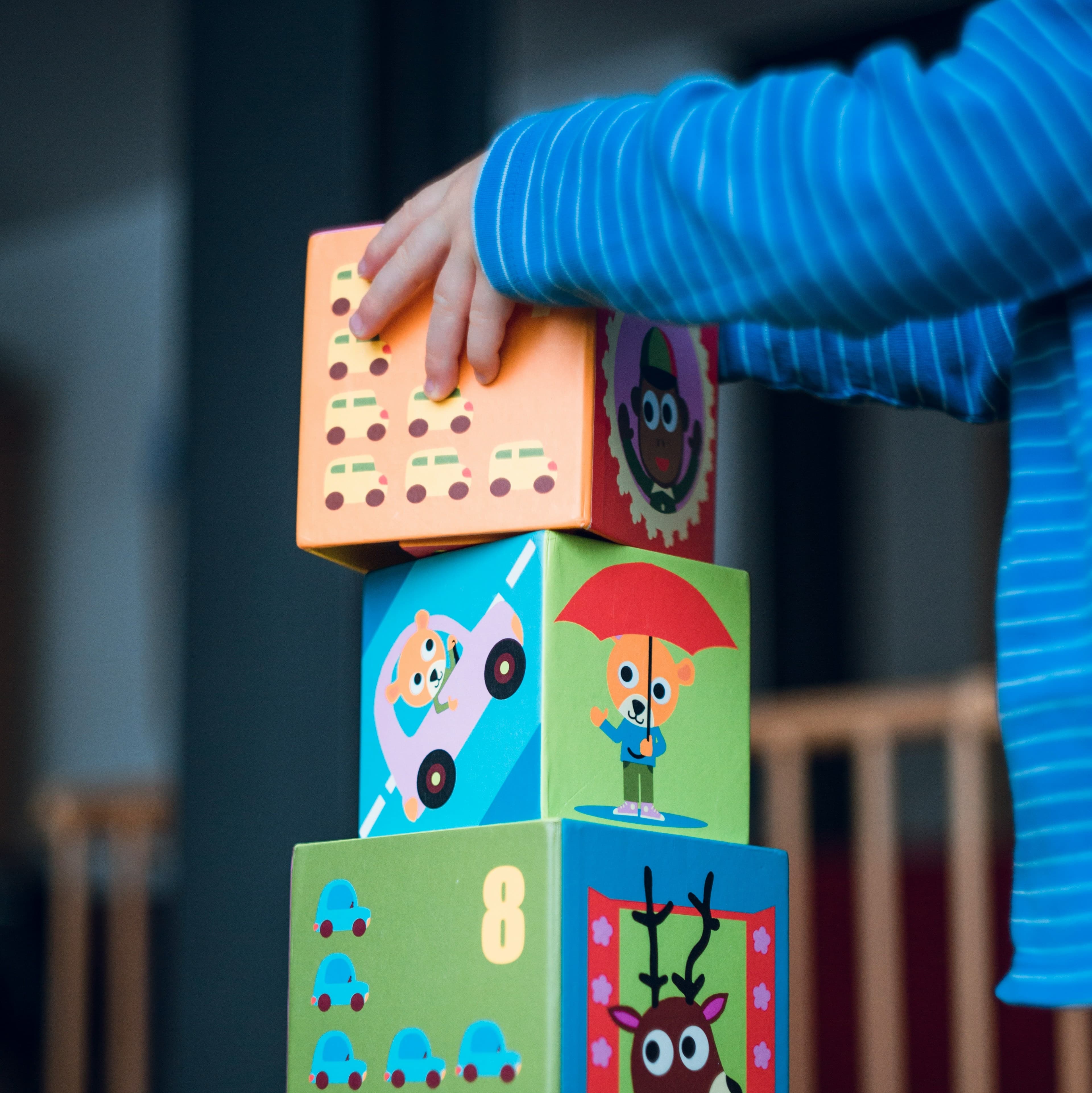 A tower of children's blocks. The blocks already on the tower come up past the waist of a child in a blue shirt who is mostly out of frame. The child's hands reach out and place a forth block at the top of the tower.