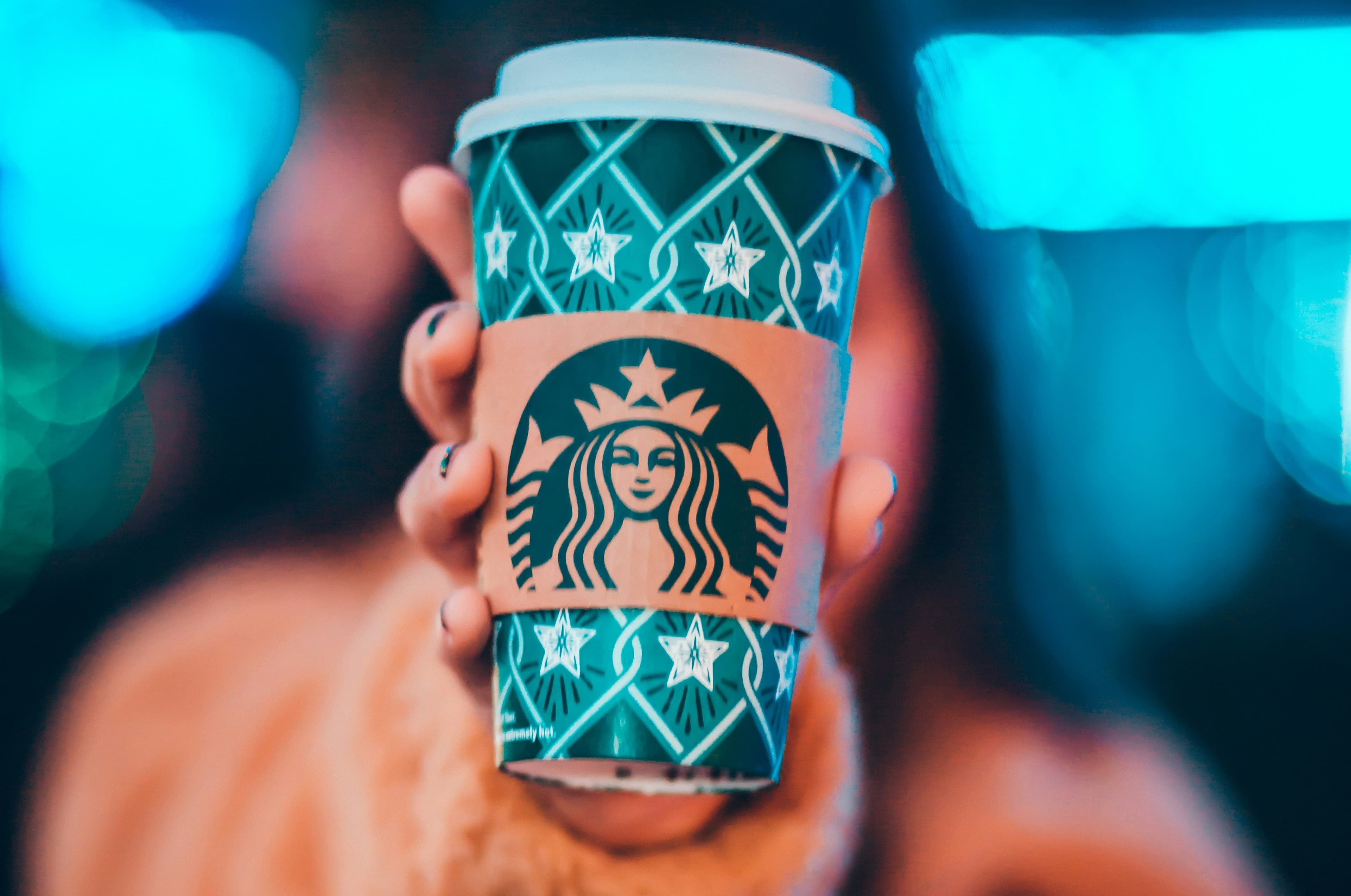 A photo of a hand holding out a blue and green Starbucks take-out cup with starts on it.