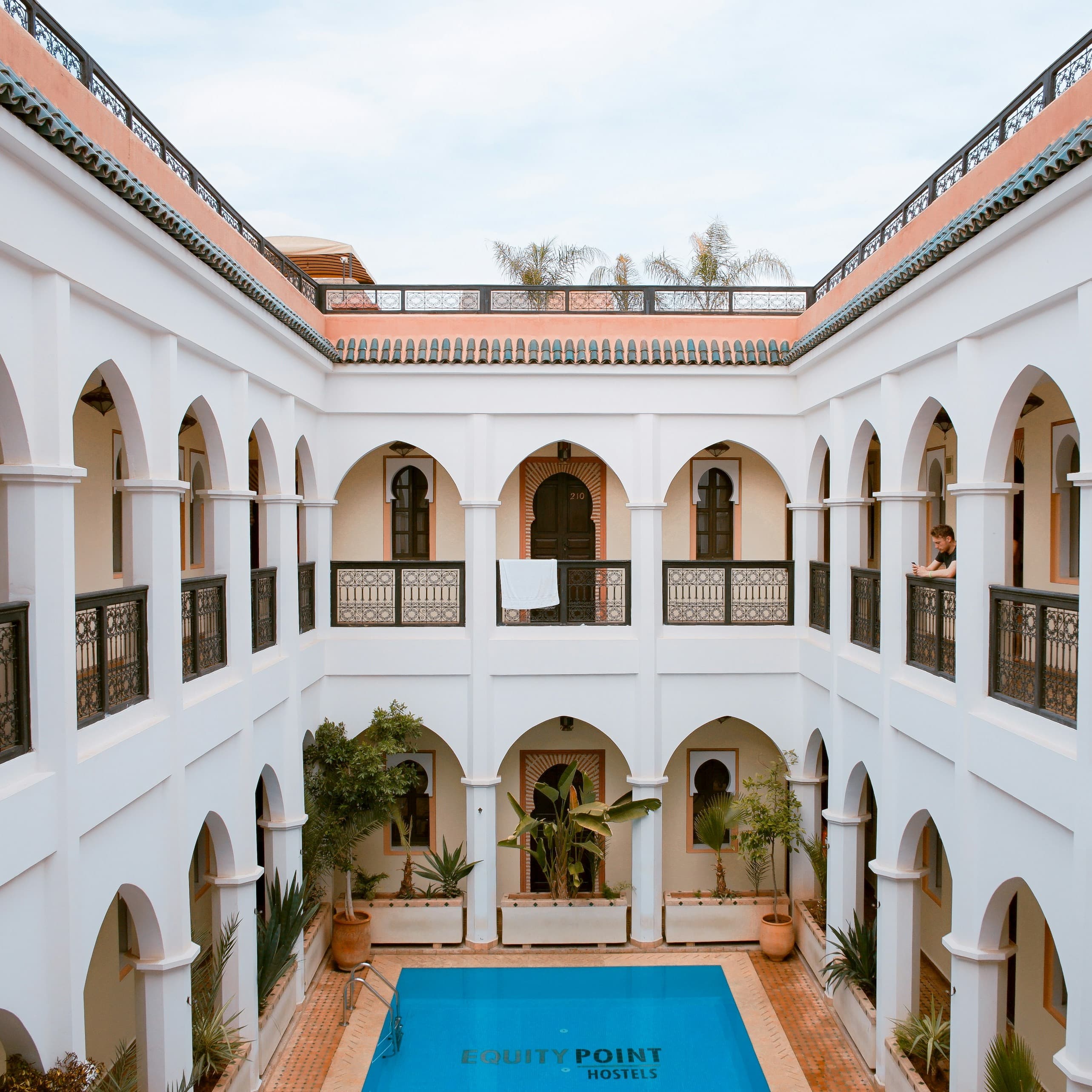 The interior courtyard of a building. Two stories of walkways surround this enclosed space. The walls of the building are white and on both stories the walkways are separated from the courtyard by North African style arches. Most of the courtyard is taken up by a very blue swimming pool surrounded by terra cotta tiling.