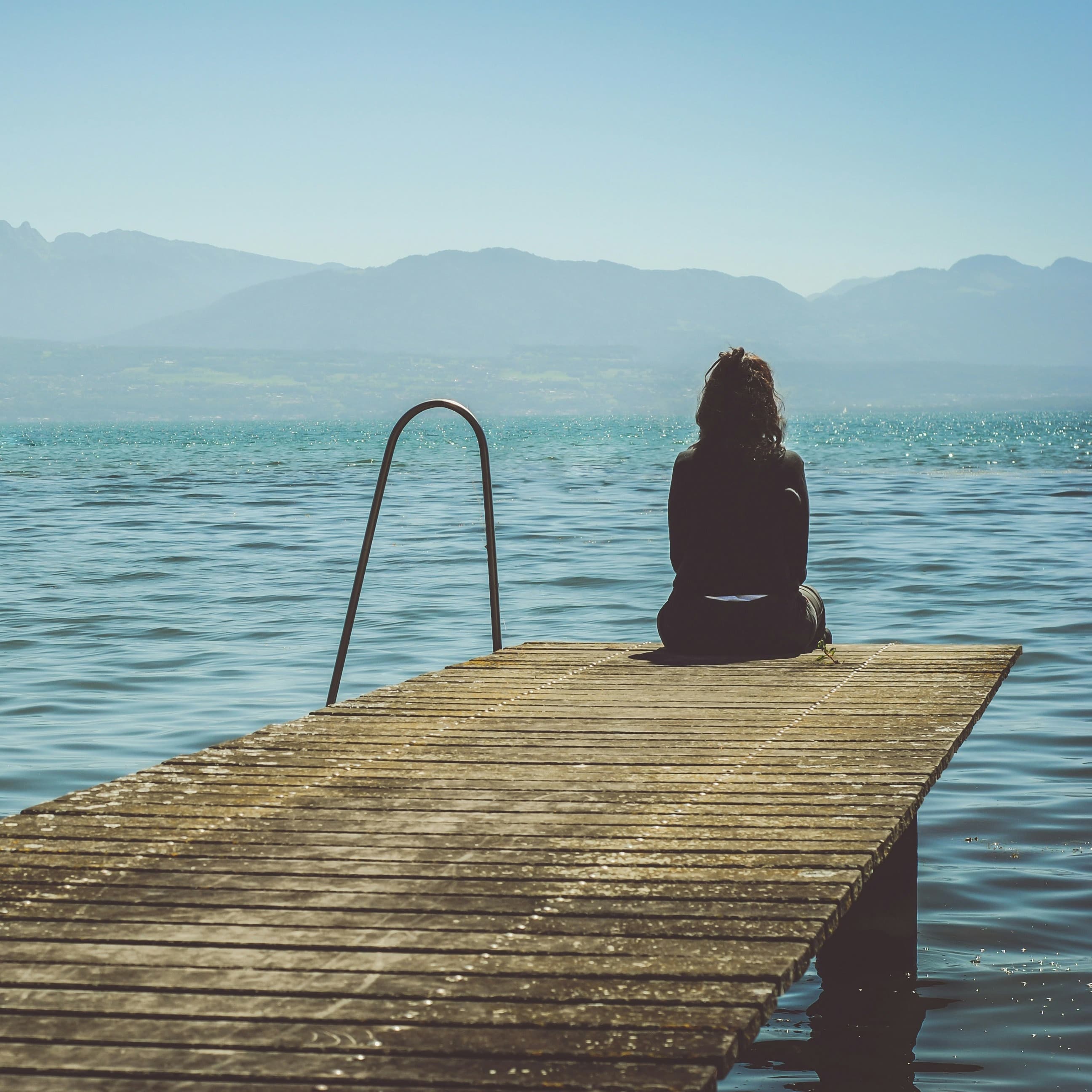 A photo of a woman sitting at the end of a dock, taken from behind. Beyond where she sits is a large lake and, on the other side, a hazy mountain rainge.