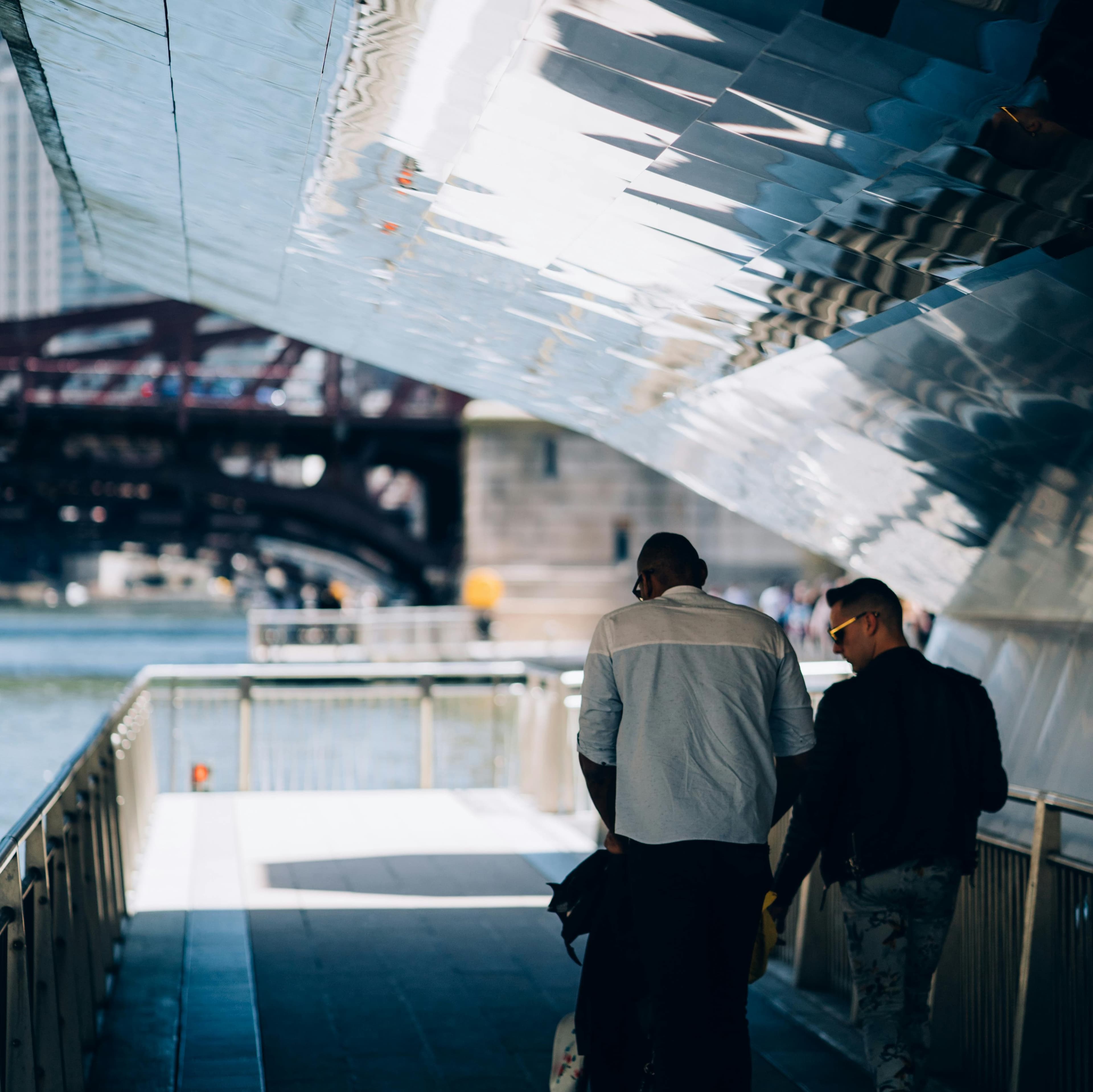 Two well-dressed men walk on a modern sidewalk. One is pushing a baby carriage. Over their heads is an arching reflective covering that leaves them in the shade. To their left is a river. Ahead of them (a little blurry), we can see an old industrial bridge and other people on the river walk.