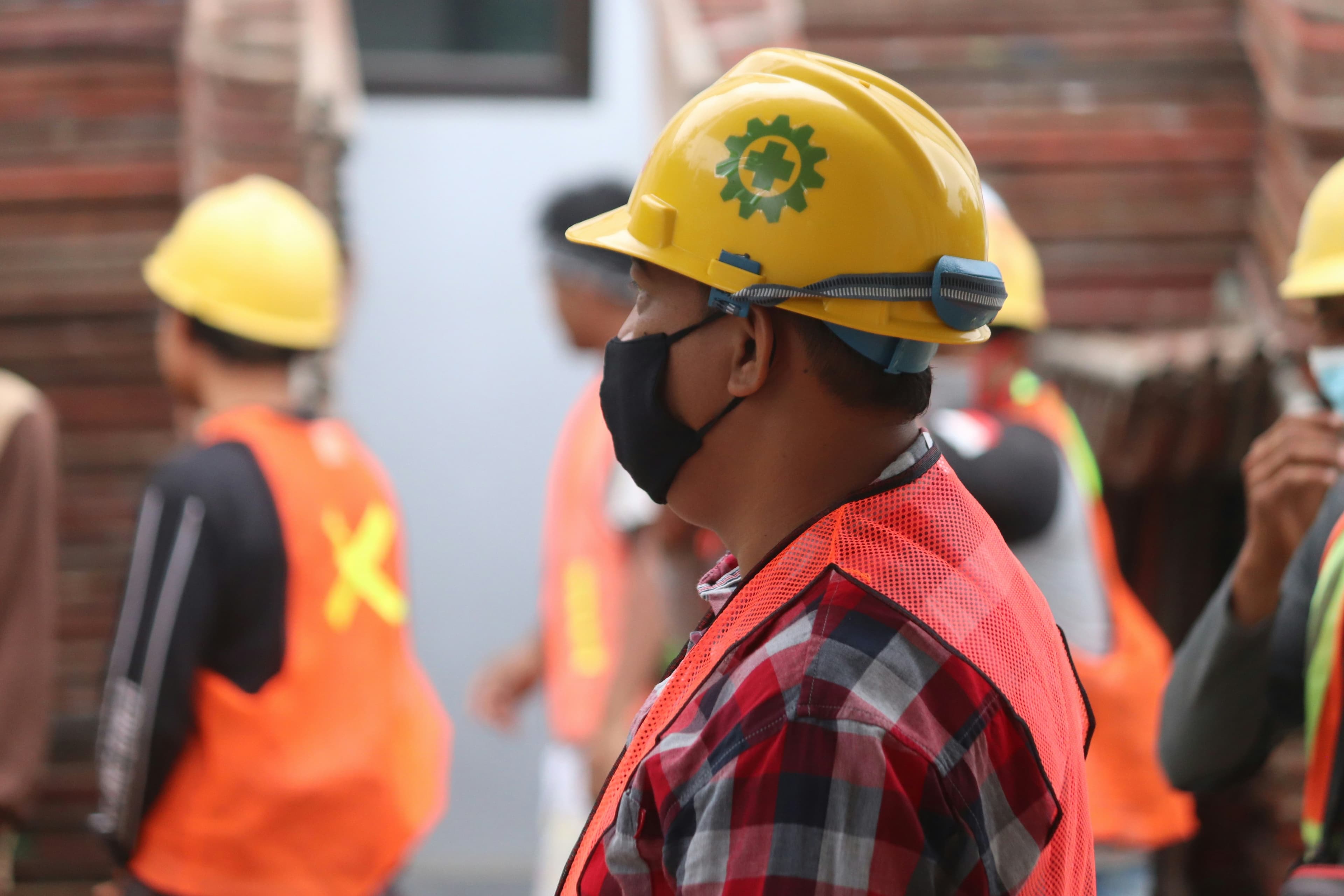 A photo of a construction worker, wearing a yellow hard hat, an orange visibility vest, and a black face mask. In the background, we can see several other people wearing the same outfit.