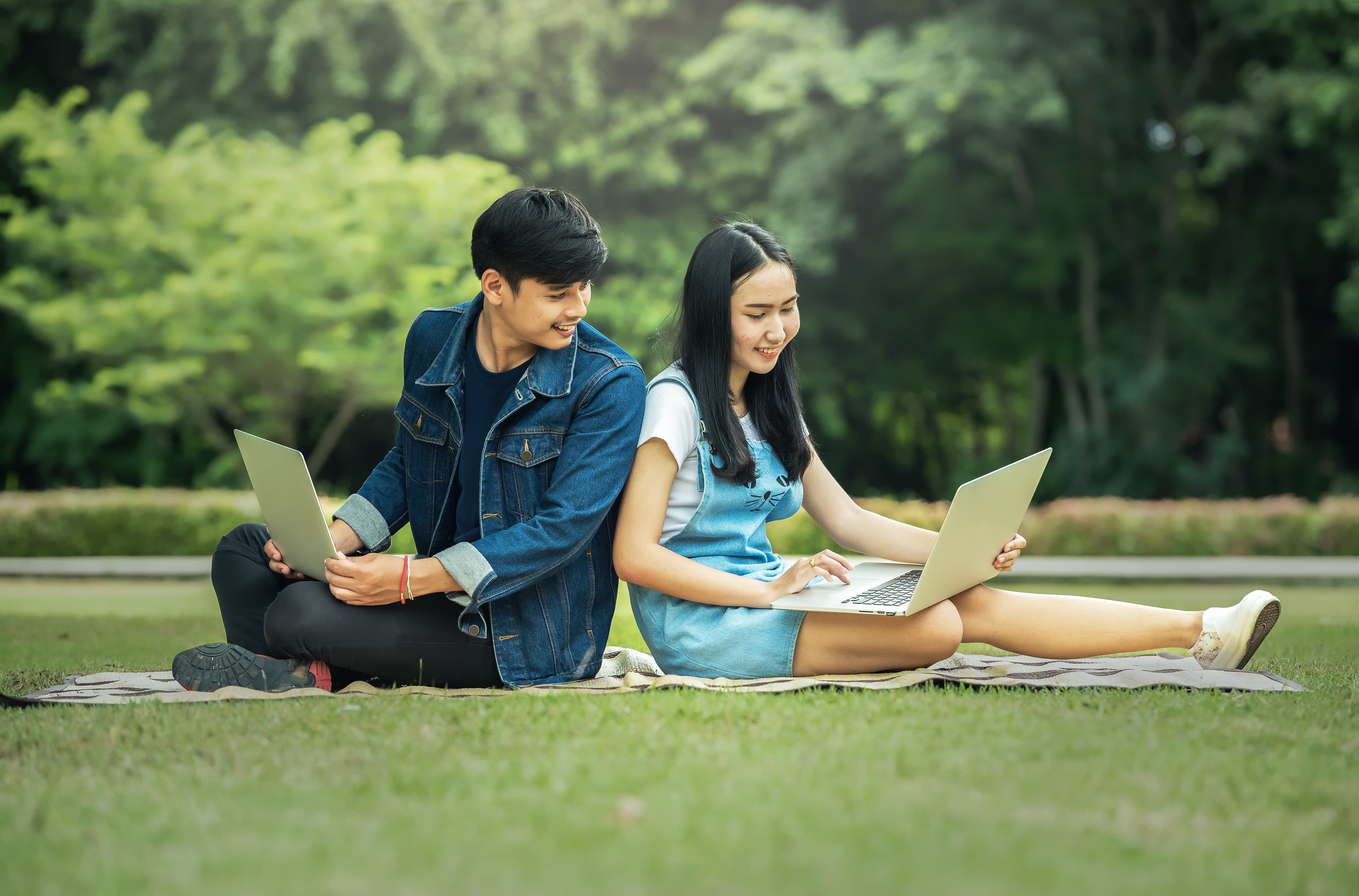 A young east Asian man and woman are sitting back-to-back on a blanket in the grass. Each has a laptop open on their lap and the man is peering over his shoulder to look at the woman's screen.