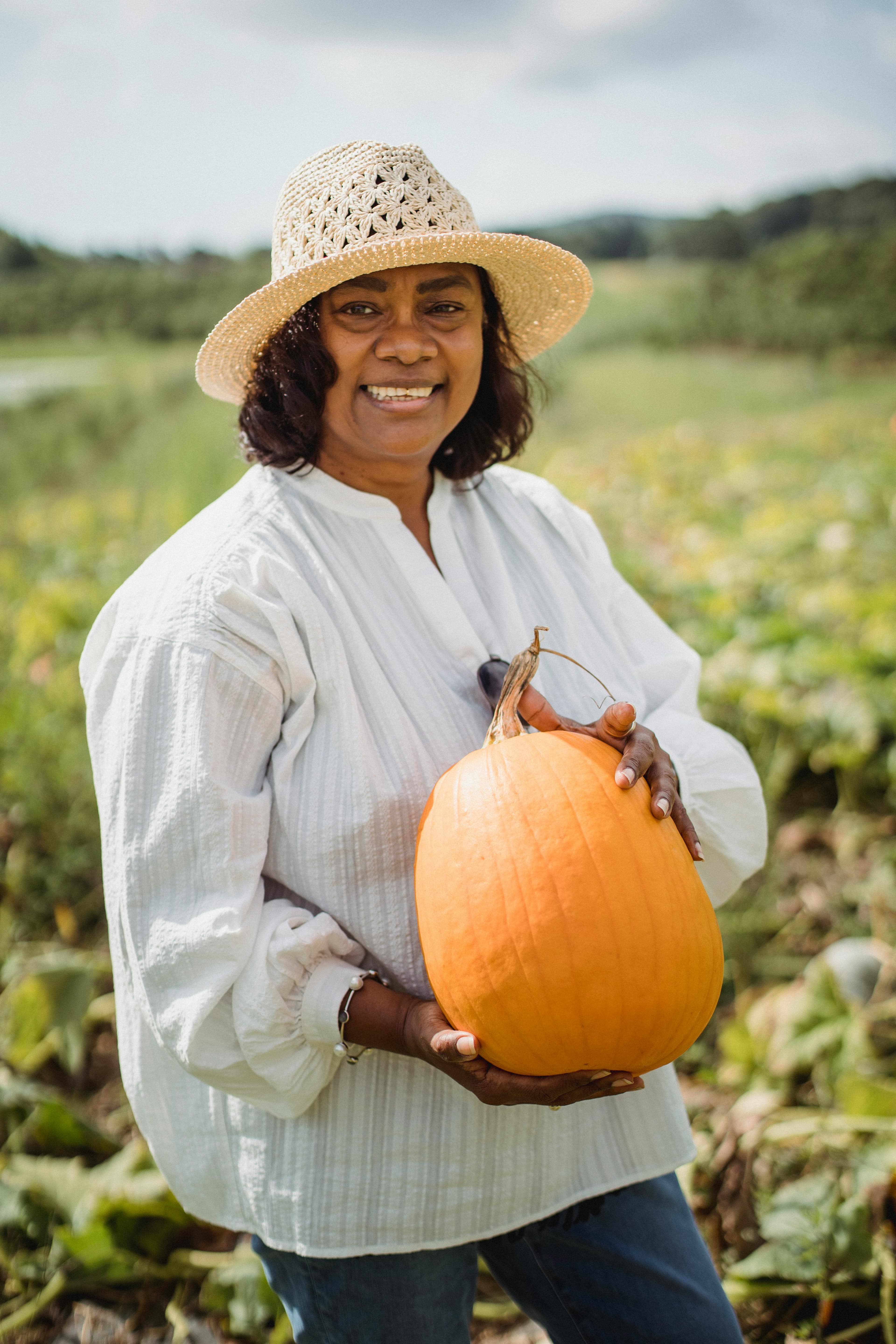 A smiling woman with brown skin wears a straw sun hat and a linen blouse and holds up a pumpkin in an agricultural field.