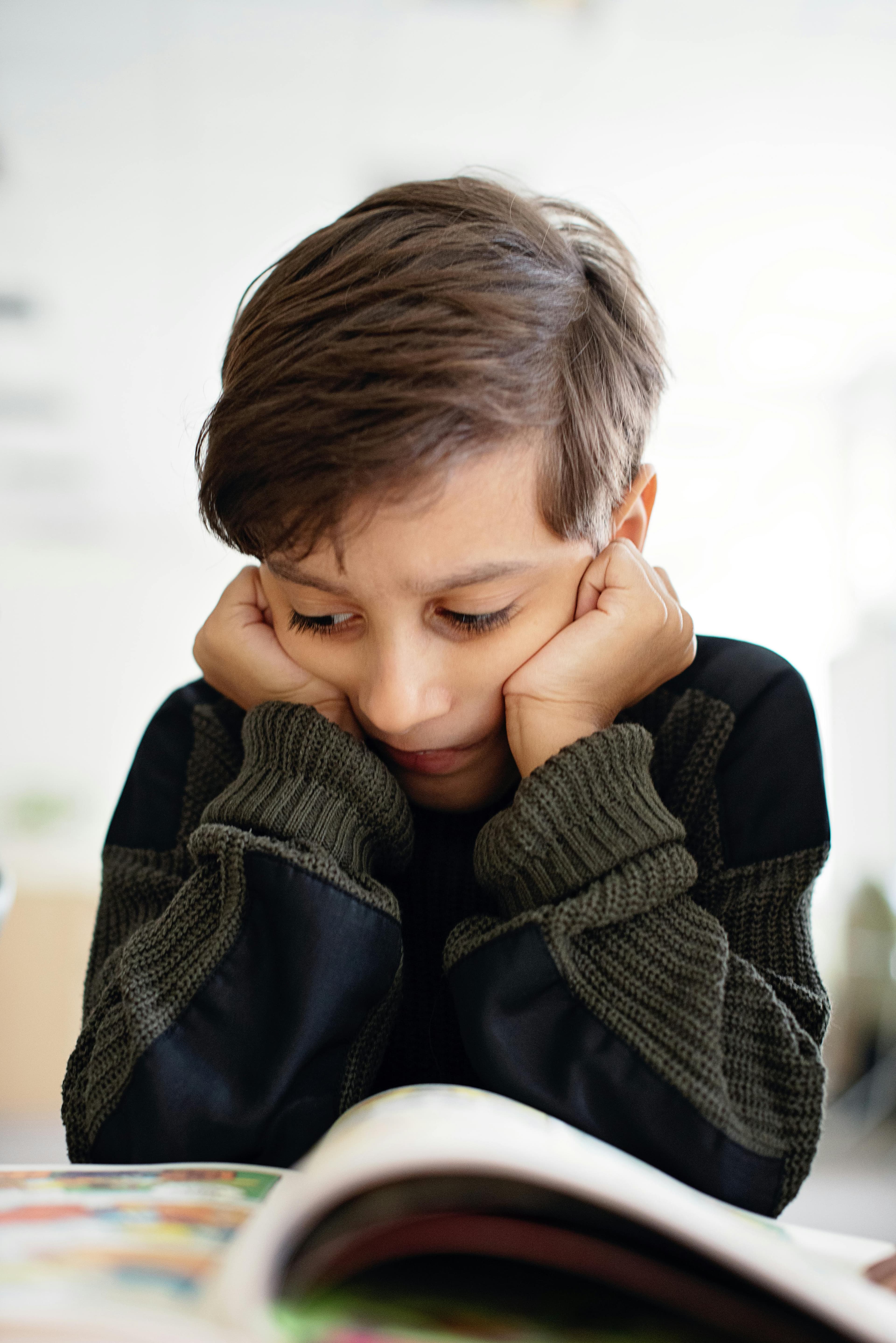 A boy with brown hair, dark eyes, and a dark sweater is holding his head in his hands while he reads a book.