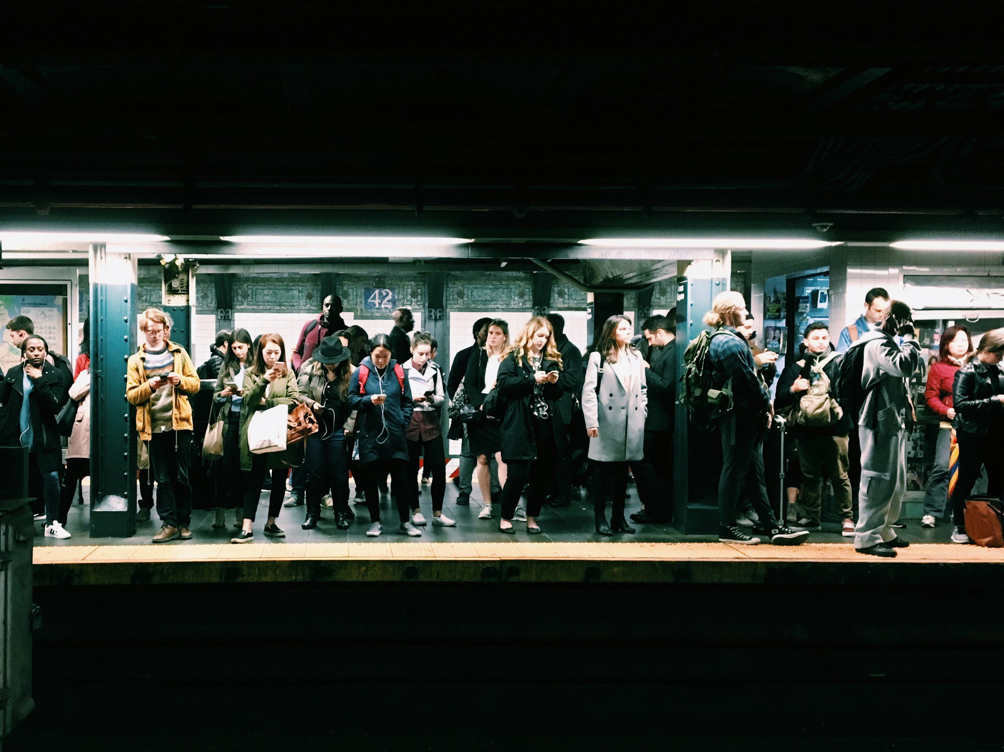A line of people waiting for a subway train. They are mostly wearing coats and almost all of them are looking down at their phones.