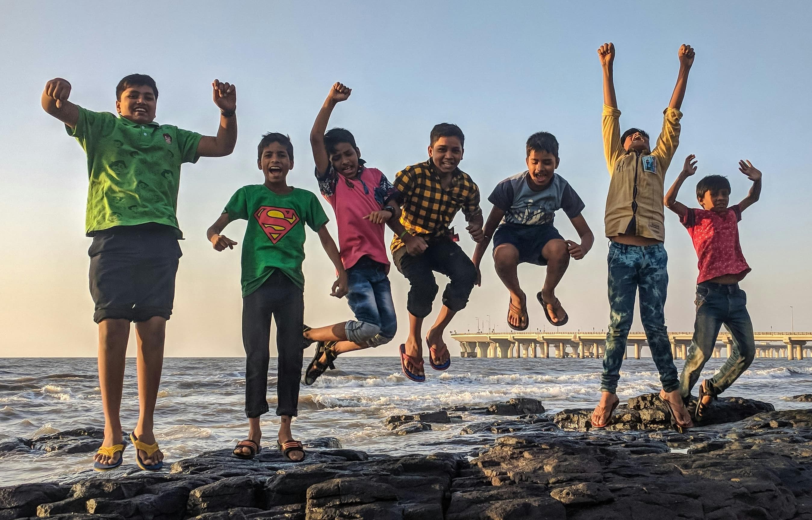 A photo of six brown-skinned boys at a rocky shoreline. They have all jumped into the air, smiling, and the picture has been taken while all their feet are off the ground. The two boys on the left wear green shirts, while the other kids wear other colors.