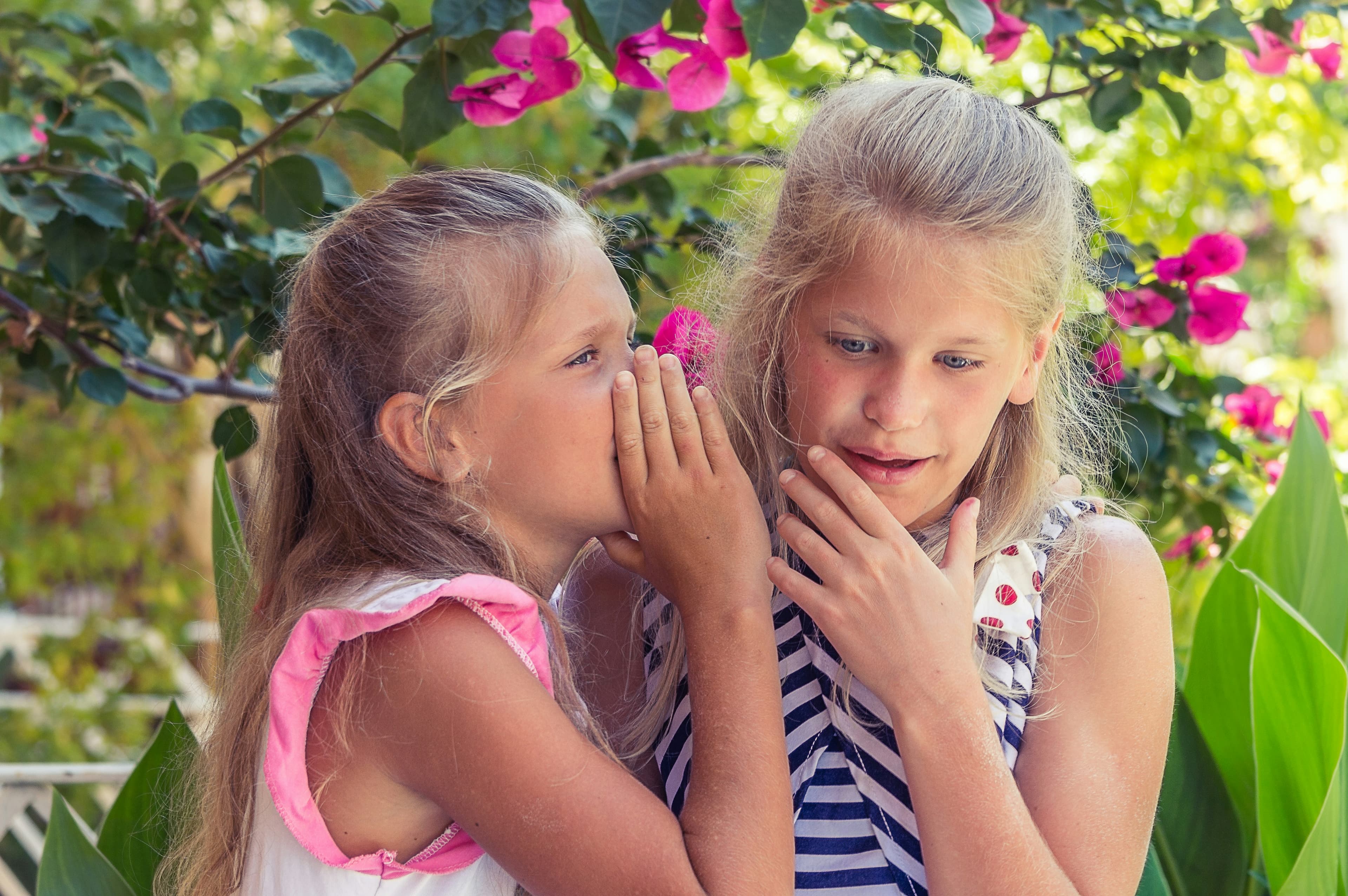 Two blond girls, about ten years old, are whispering to each other in a garden.