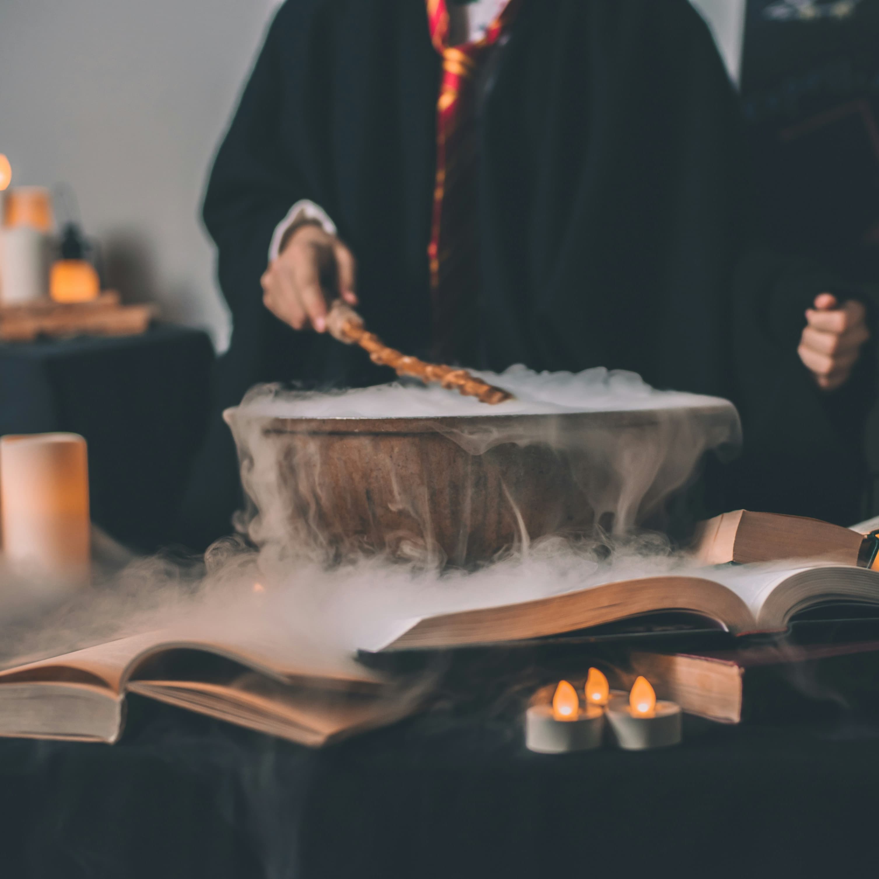A photo of a bubbling cauldron, open spell books, and faux candles. In the background someone dressed in a Harry Potter outfit pokes the cauldron with a wand.