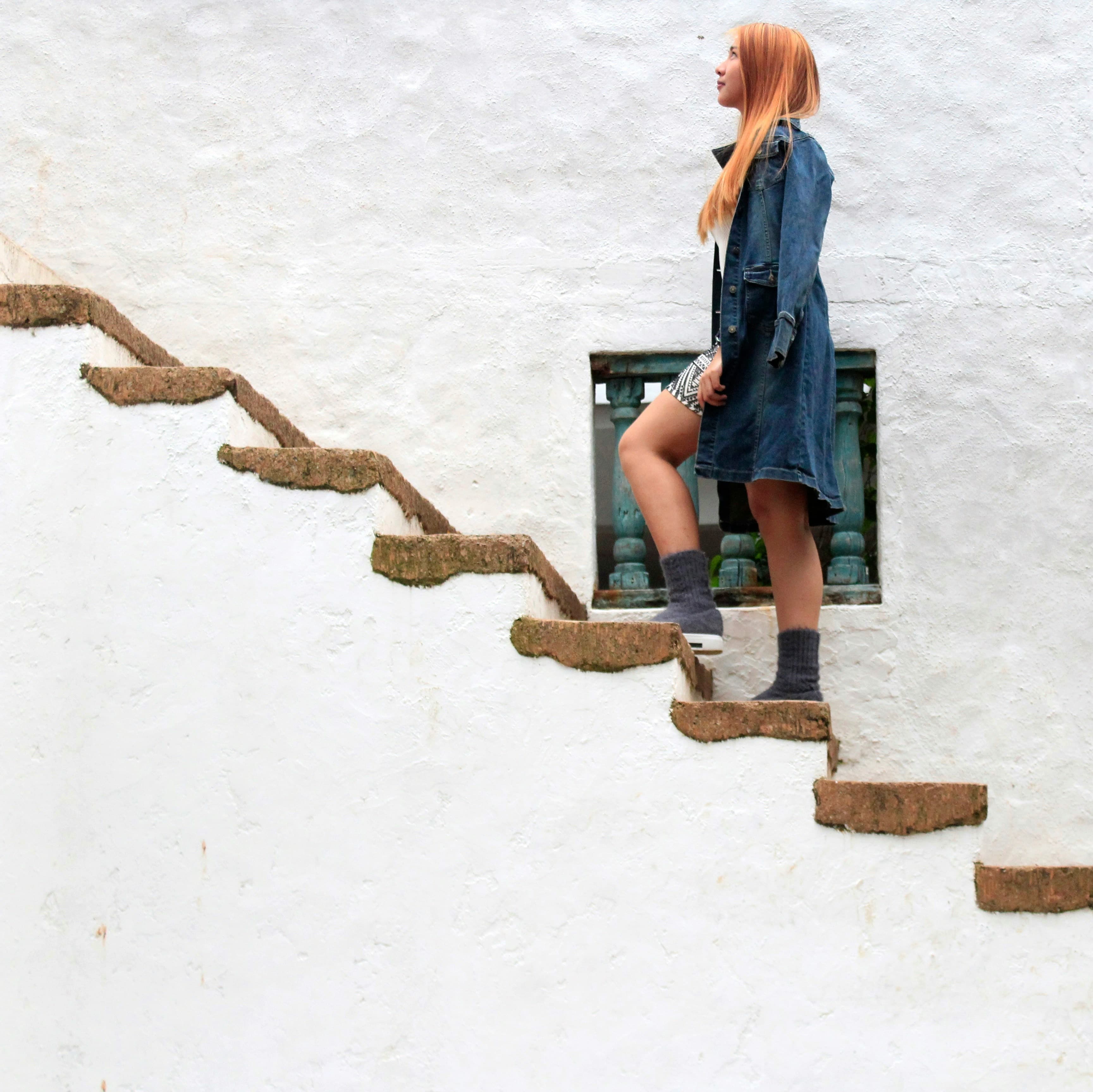 A photo of a woman with long orange hair walking up some stairs. The photo is take from the side, so we can see the side of the stairs and the wall behind them. Both are lime washed white, there is no railing, and the treads on the stairs are made of wood.