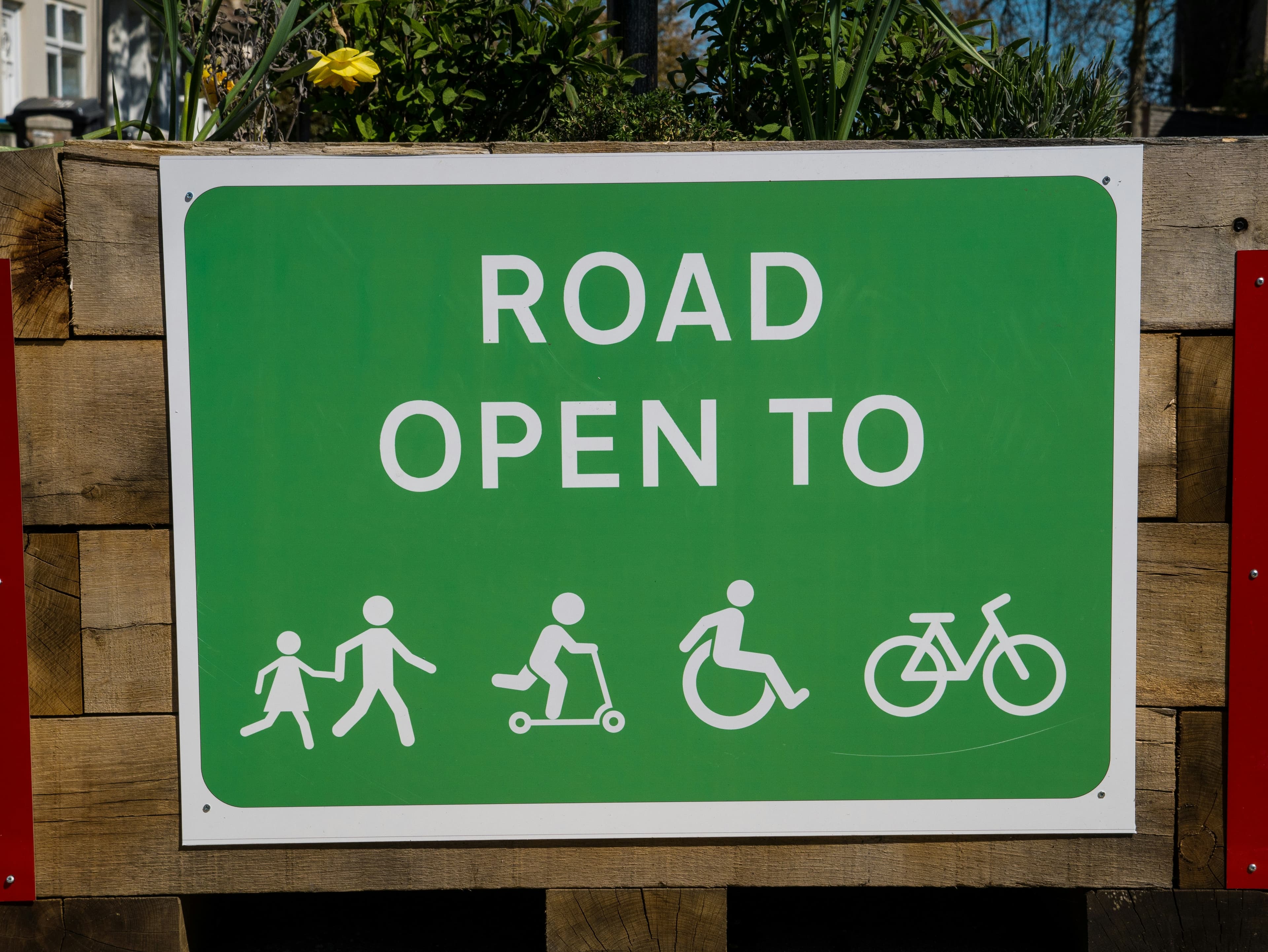 A green street sign reading "road open to." Underneath, this a graphic of a bike, a person in a wheelchair, a person on a scooter, and two people running hand-in-hand.