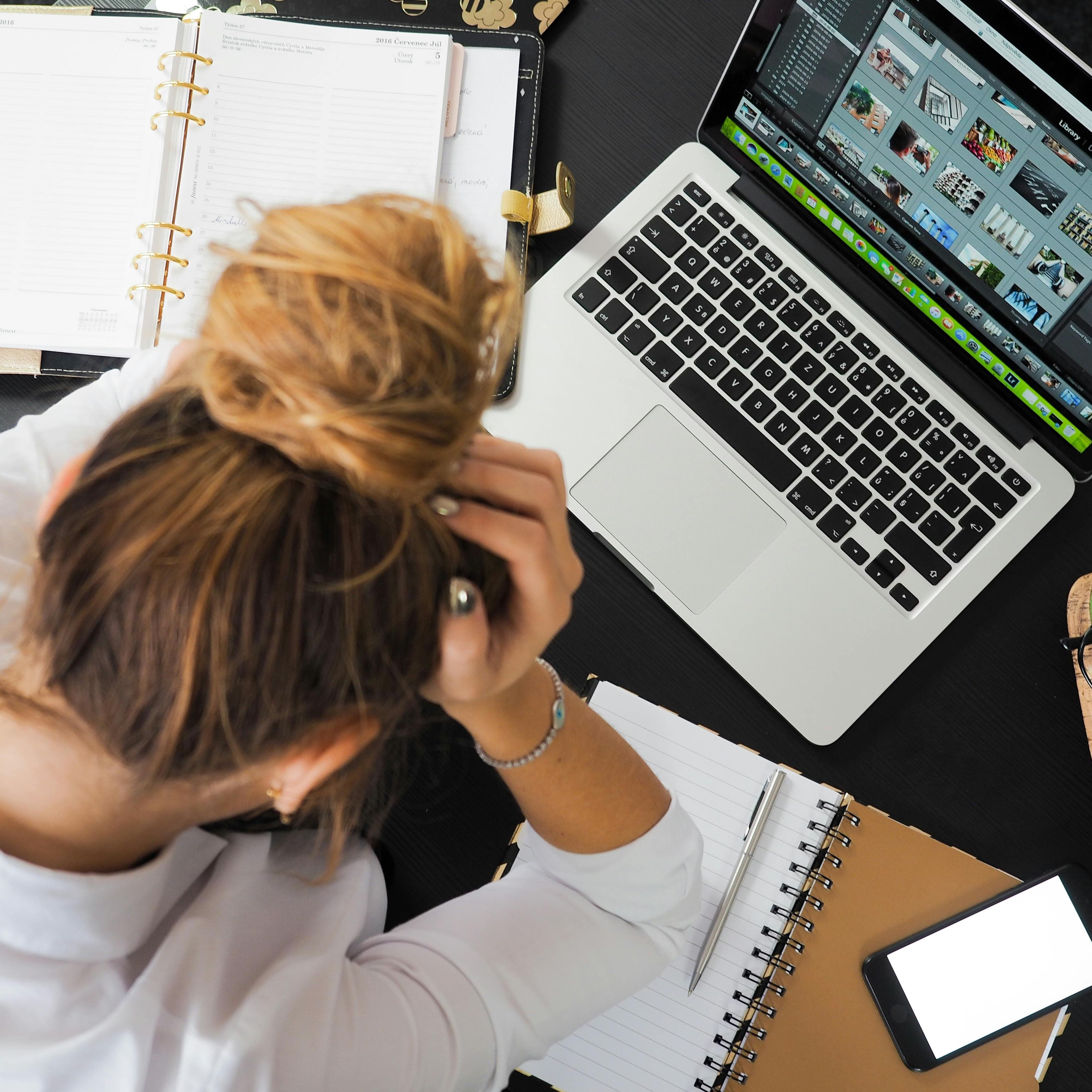 A photo of a woman taken from above. She has a planner, a notebook, a phone, some glasses, and a laptop open on her desk and she is holding her head in her hands, clearly overwhelmed.