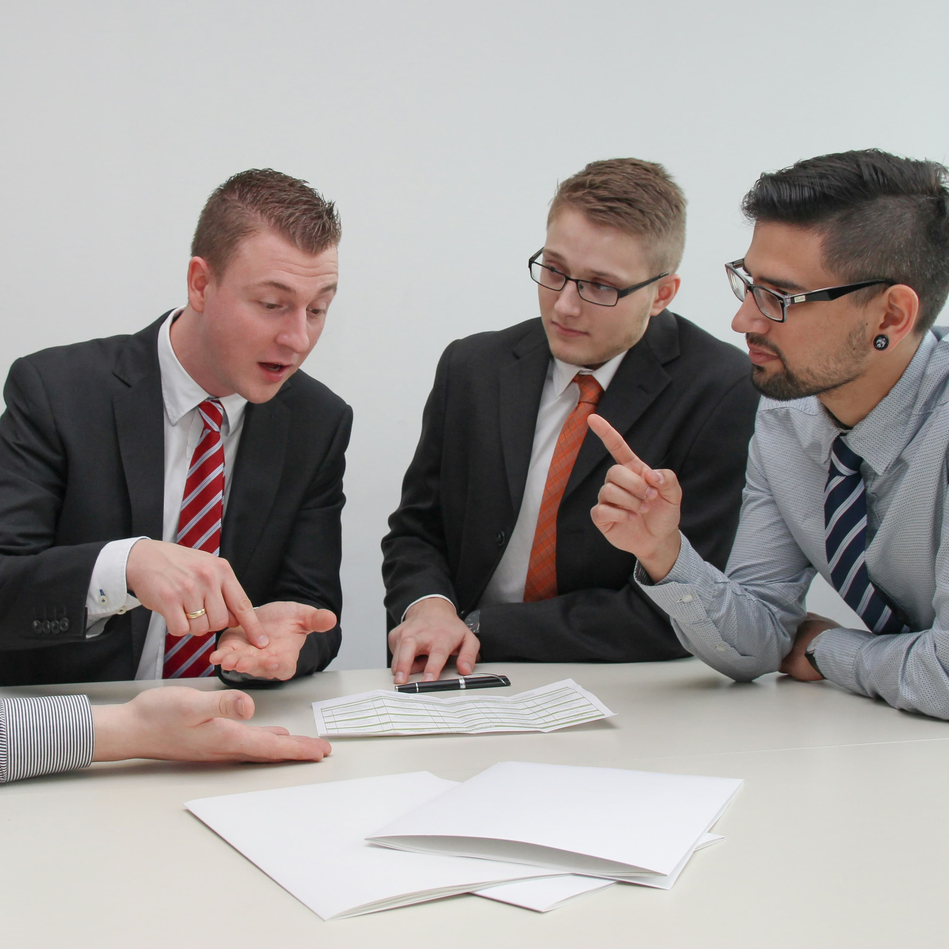 A group of white men in business outfits are sitting around a white table. They are having a debate about the contents of a sheet of paper that sits between them.
