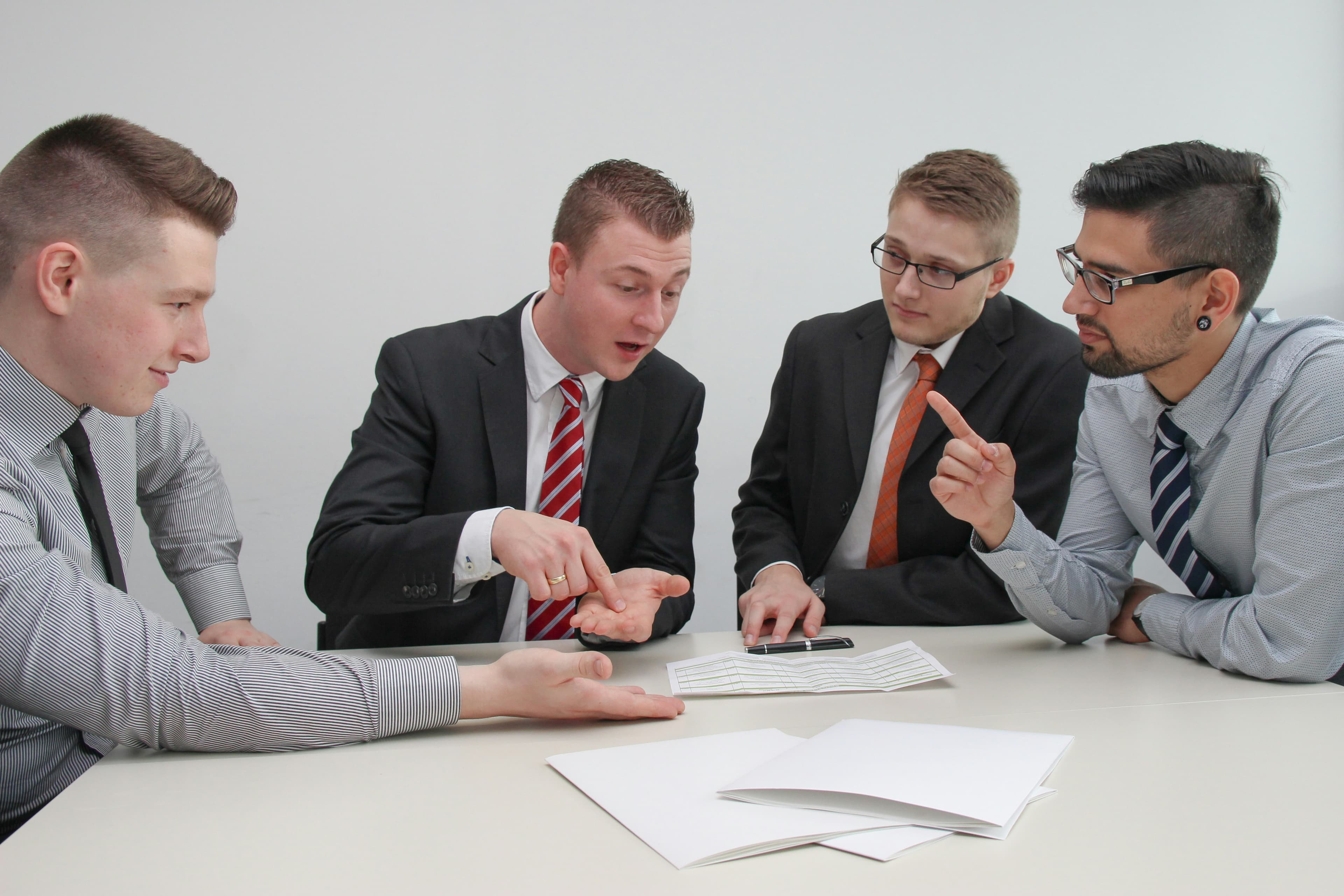 A group of four men in business attire sitting around a table and discussing some papers.