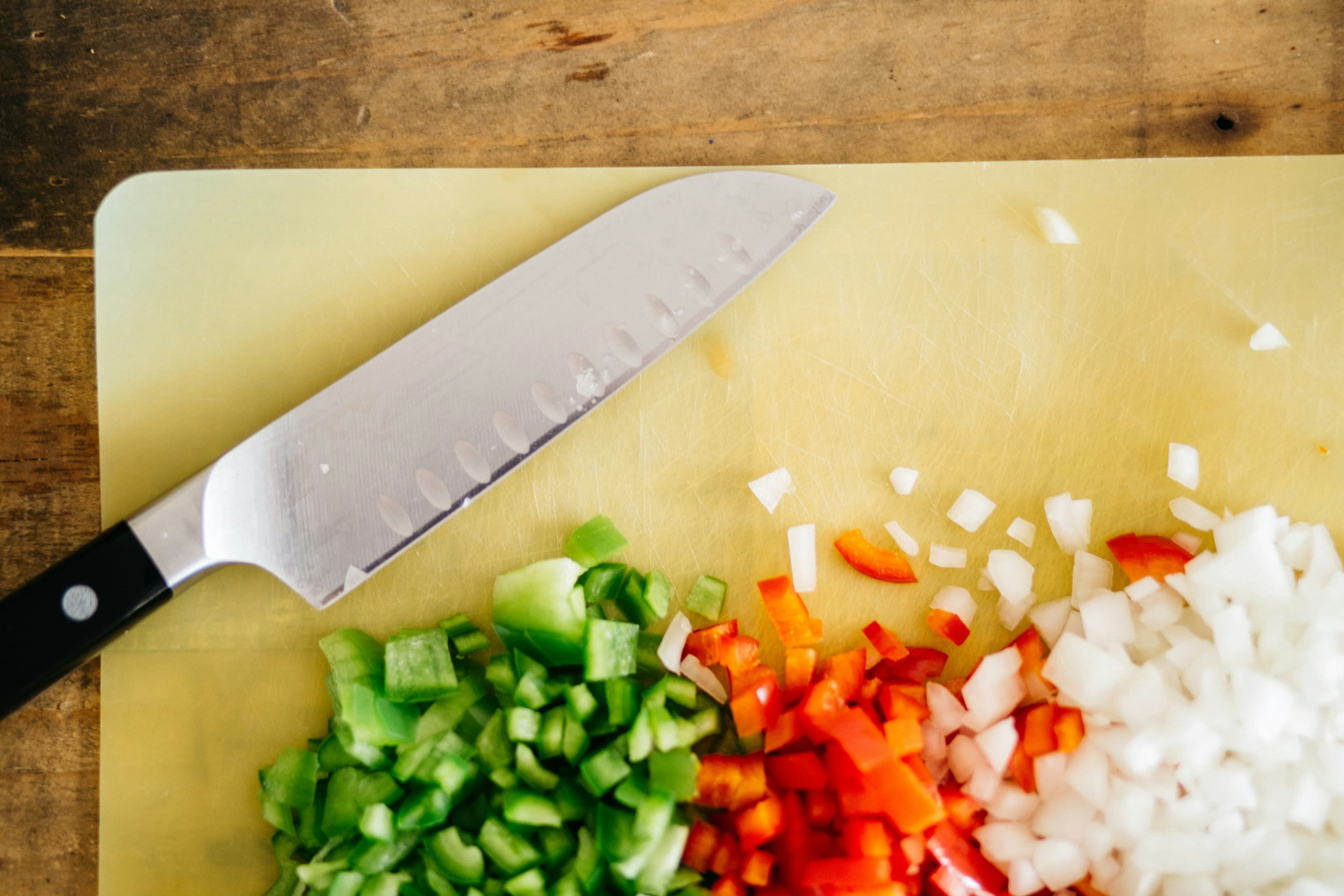 A kitchen knife on a yellow cutting board with some diced vegetables (peppers and onions)