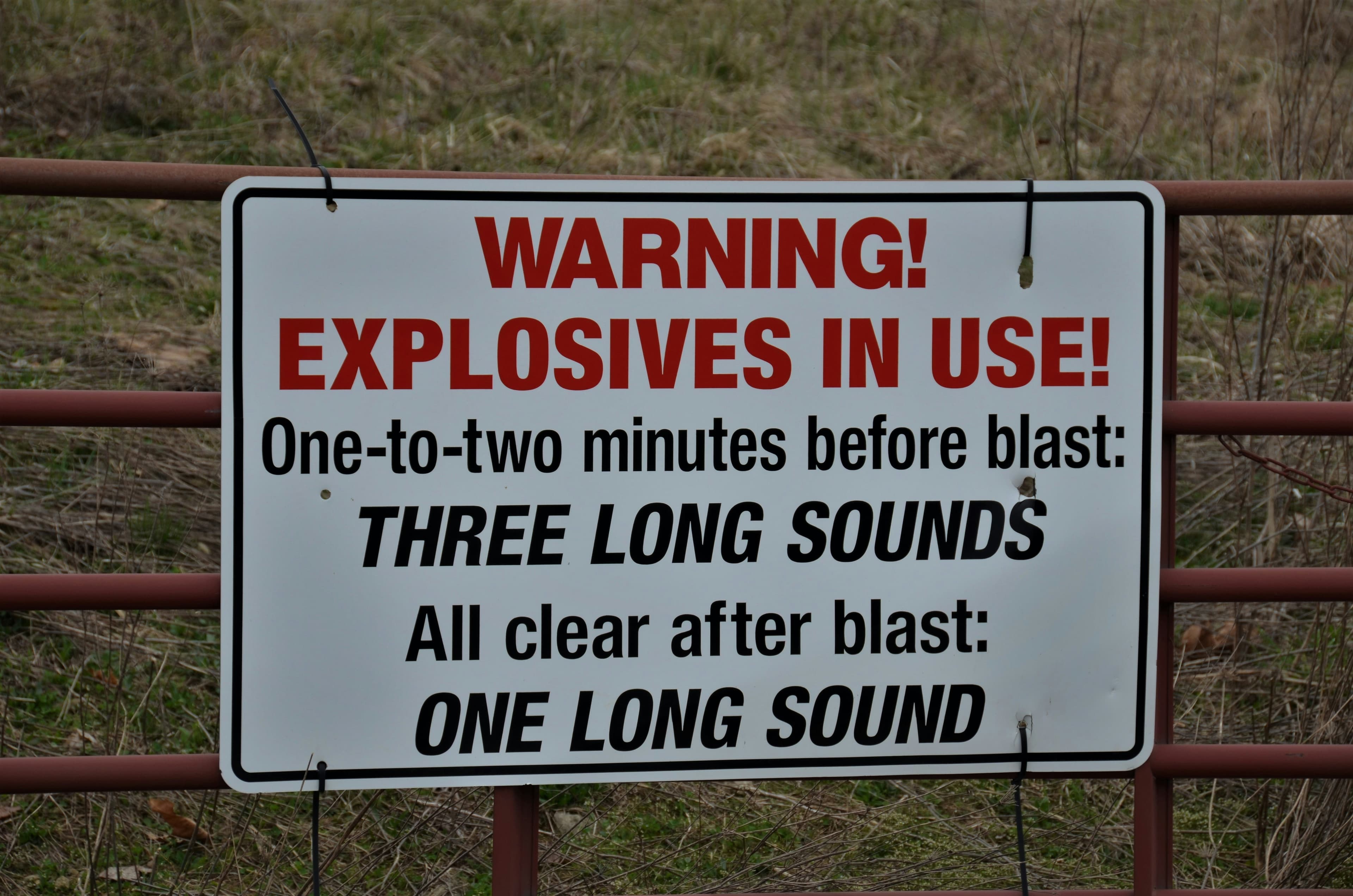 A white rectangular sign. A red heading says "Warning! Explosives in use!" and the rest of the sign (black) reads "One-to-two minutes before blast: THREE LONG SOUNDS. All clear after blast: ONE LONG SOUND."