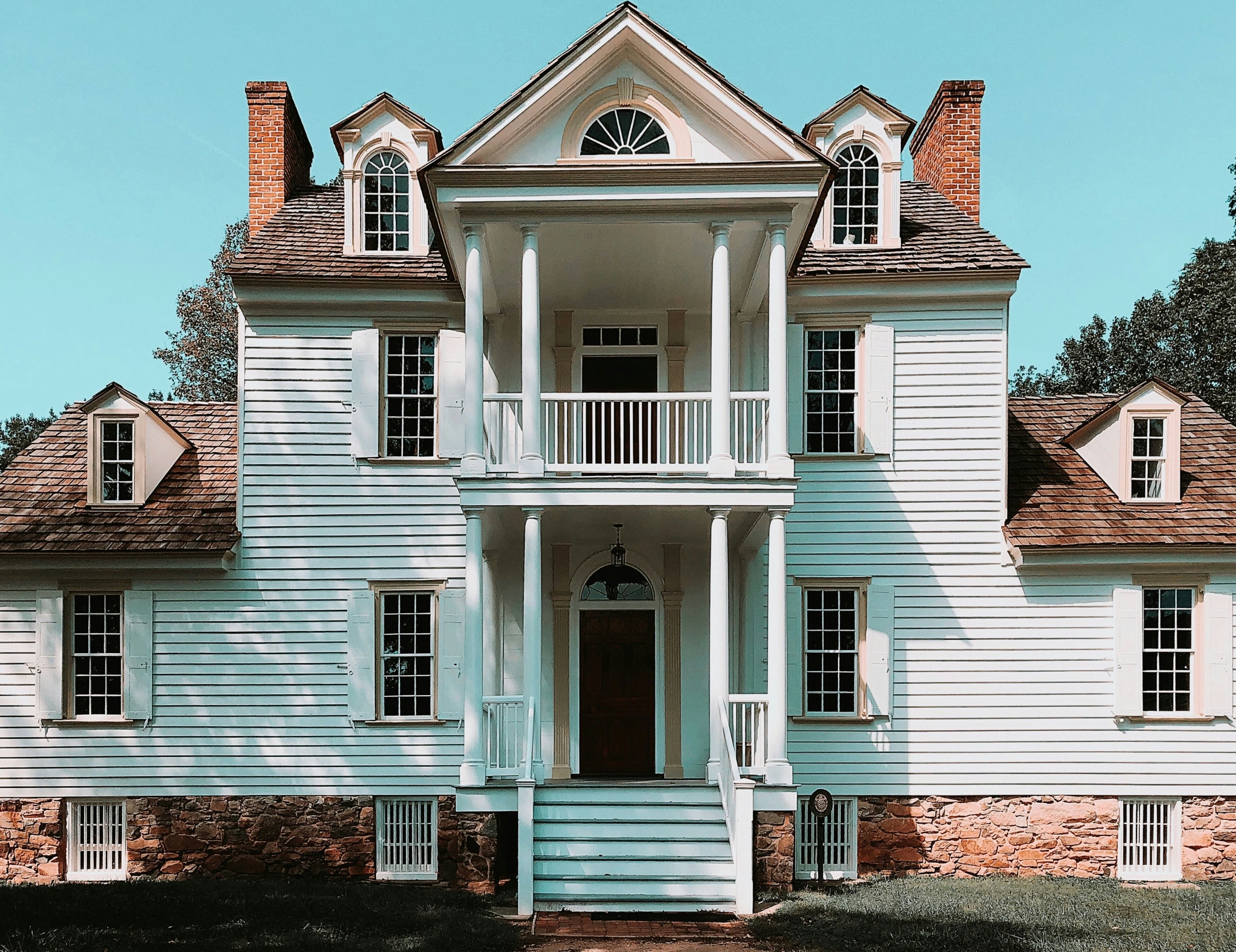 A photograph of an antique white colonial-style mansion, with a two-story columned porch and a flagstone foundation.