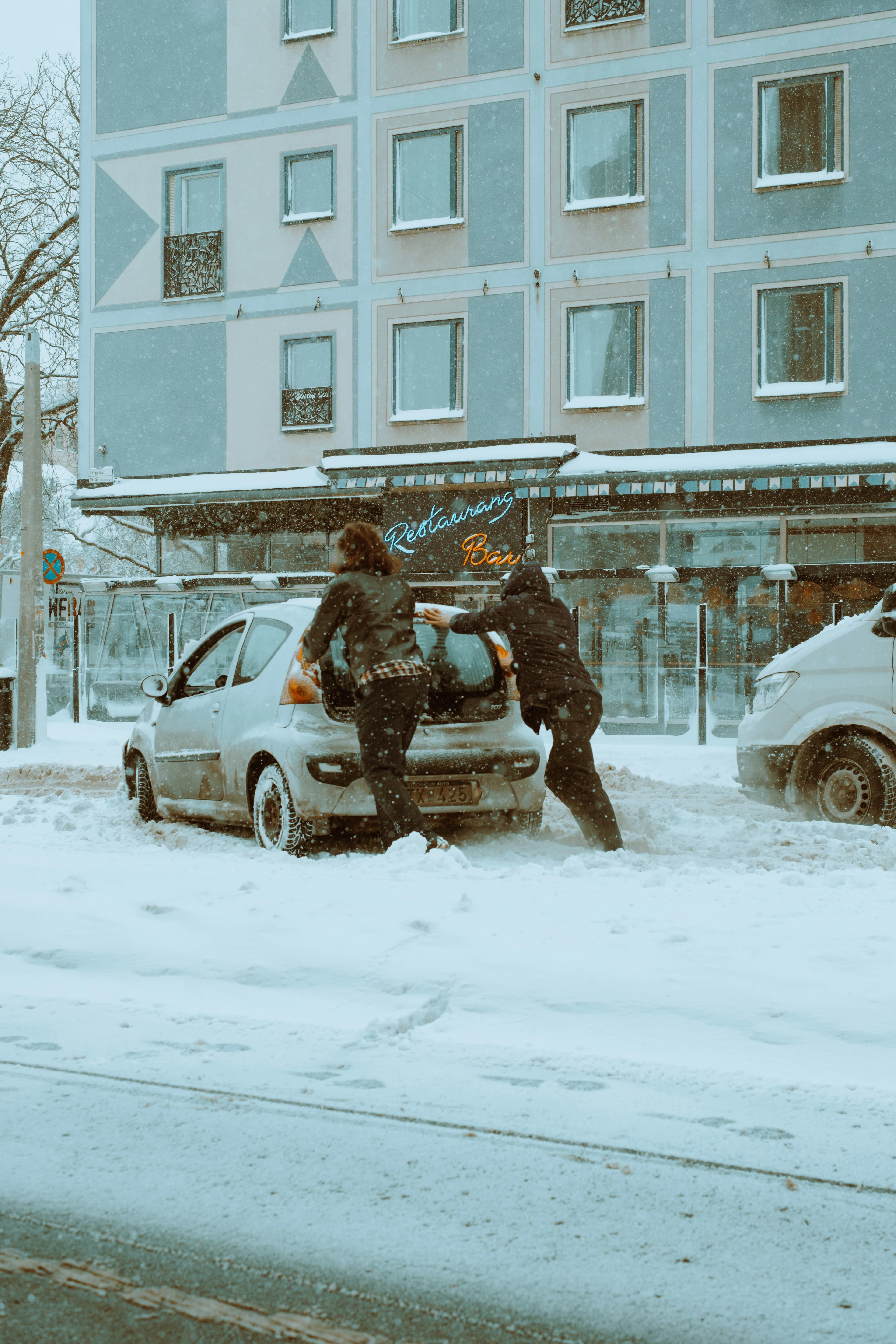 A photo of two people pushing a small dirty car down a street in a snowstorm.