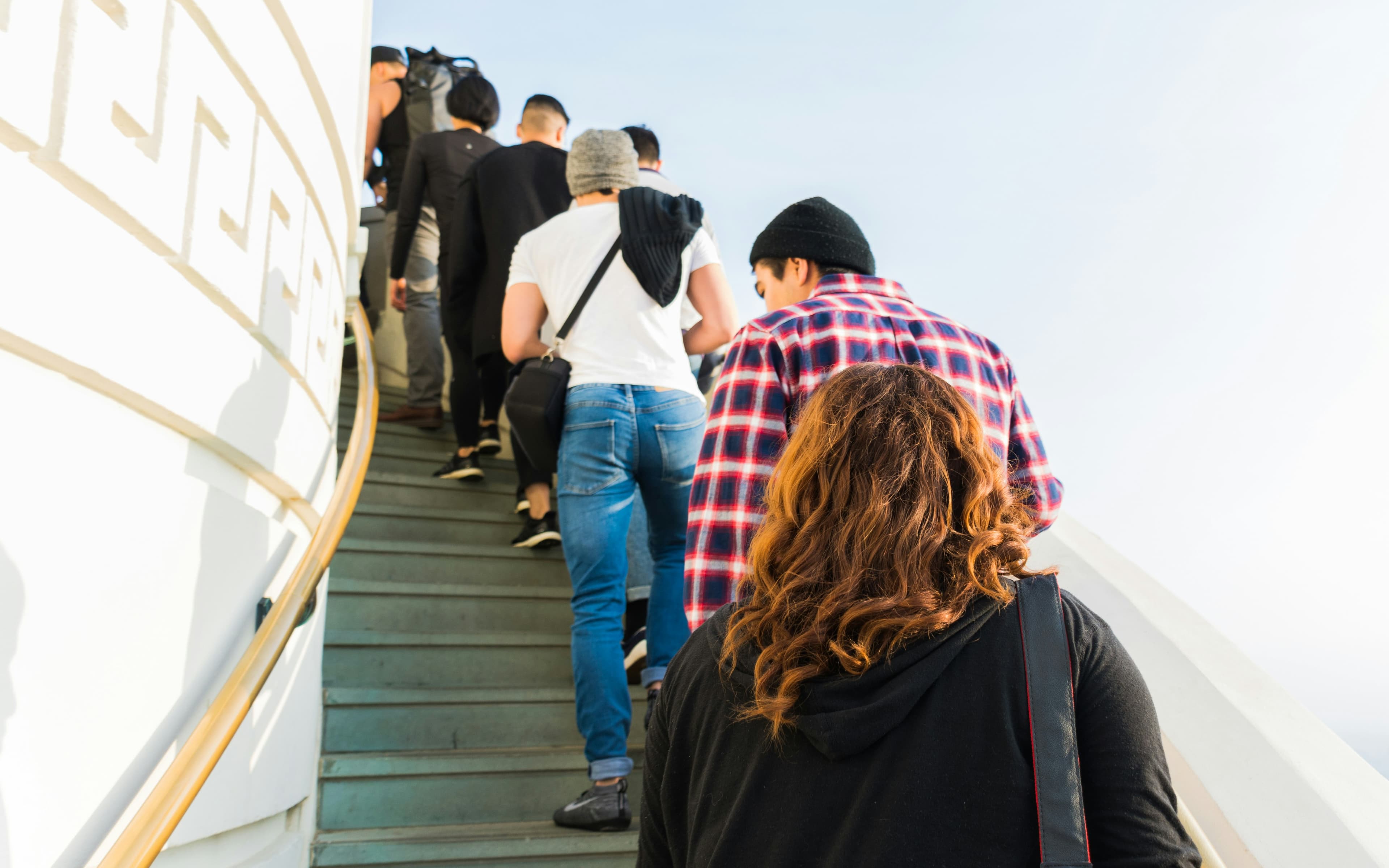 A photo of a line of people walking up a staircase. We are looking at their backs, and the staircase is curving around the outer wall of some sort of tower. The wall is white and decorated with a Greek key pattern.