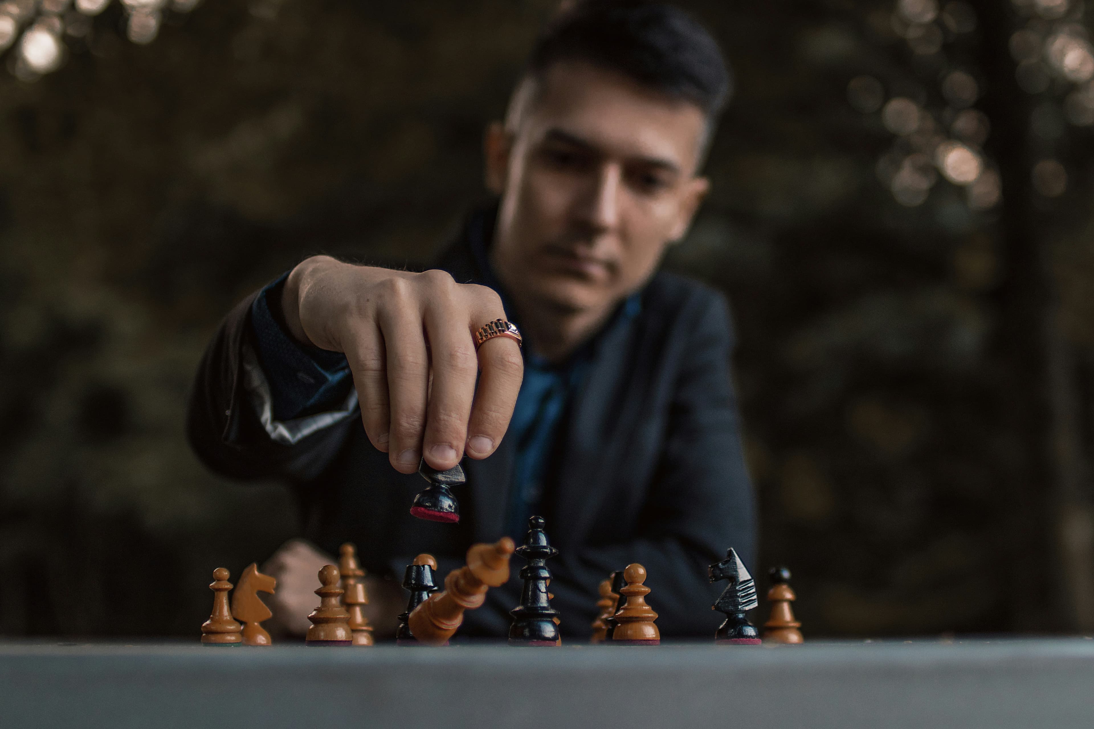 A close-up photo of a man's hand using a black chess piece to knock over the white king. We can see the owner of the hand blurred out in the background, a white man with short dark hair playing chess outside.