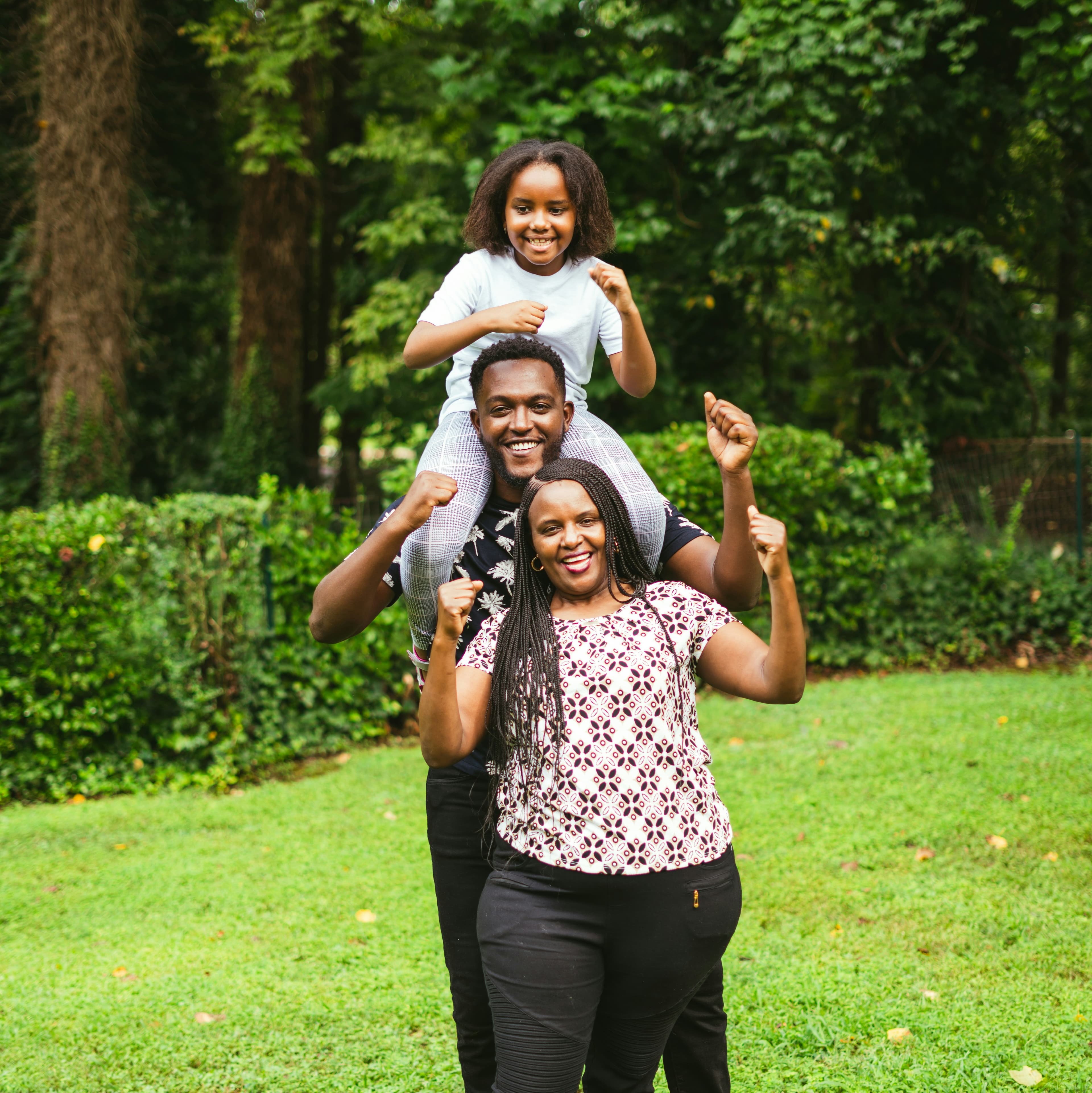 A photograph of a family of three stands in a garden with their thumbs up. A woman is in front, a man stands behind her, and a little girl is on the man's shoulders.