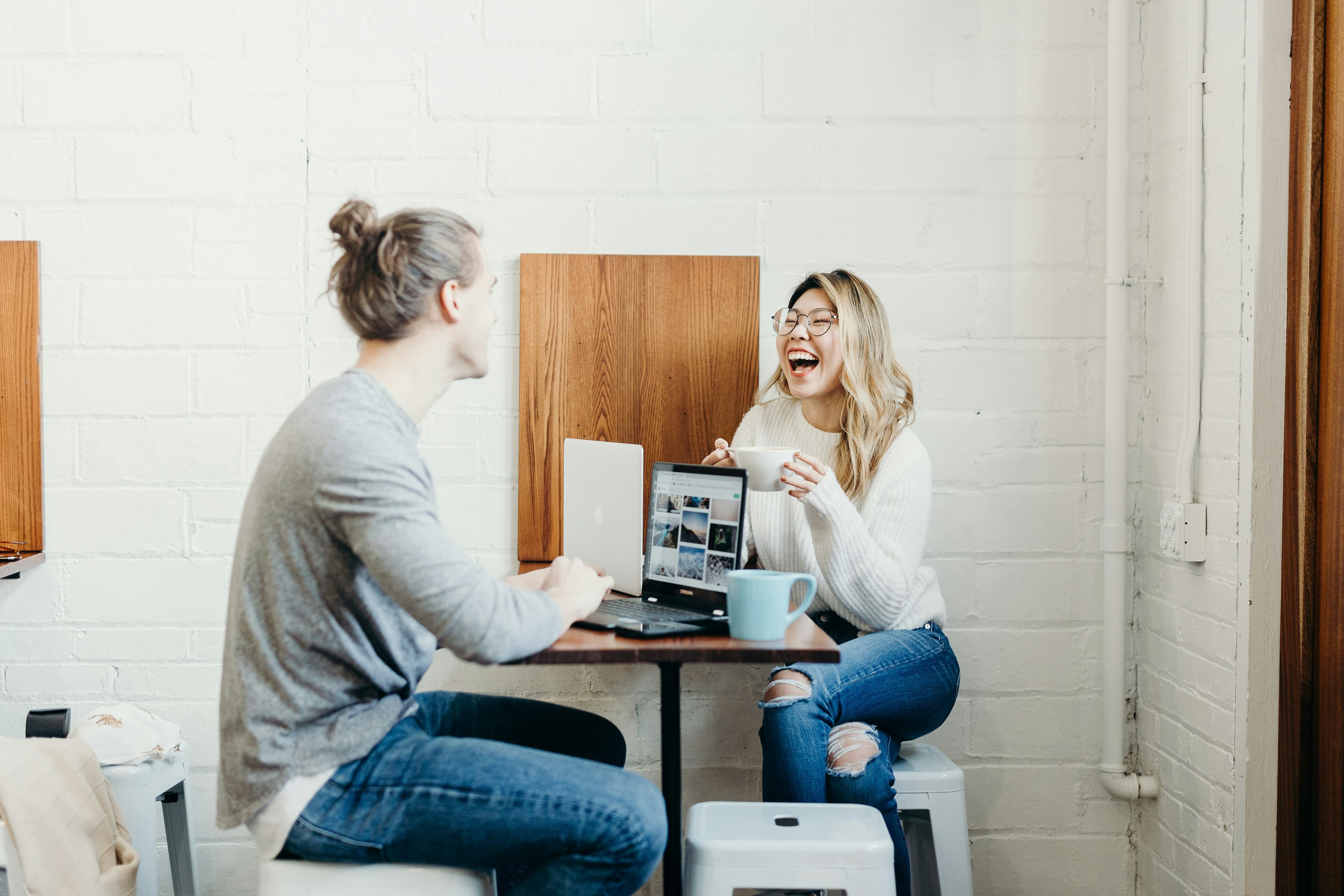 A photo of two people chatting at a cafe table, a white man with a bun and an East Asian woman with blonde highlights and glasses. The woman is laughing at something the man said and is holding a coffee mug. The man is talking with his laptop open