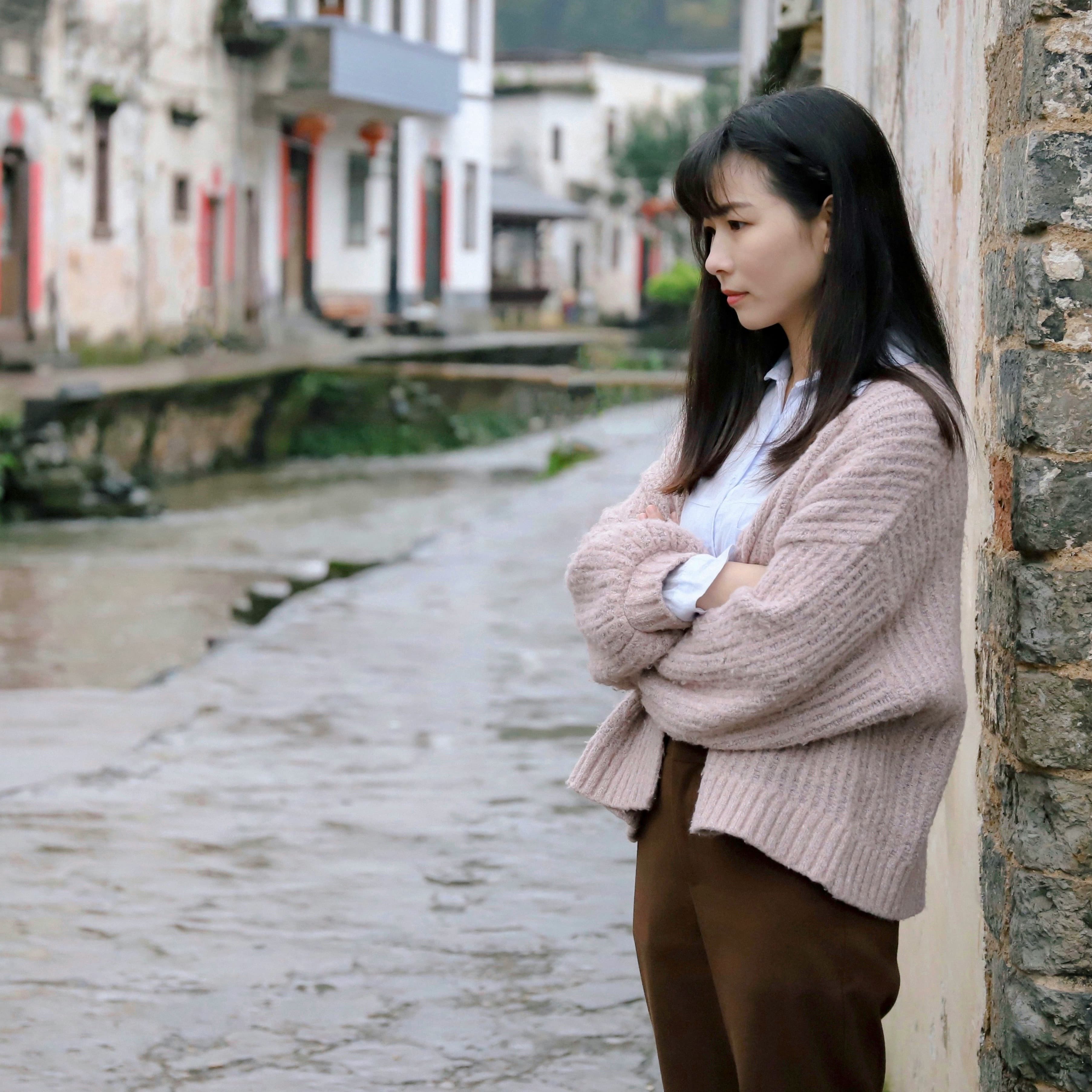 A photo of a sad woman with long black hair in a beige sweater. She stands by a wall with her arms crossed. Behind her we can see a historic looking street and a canal.