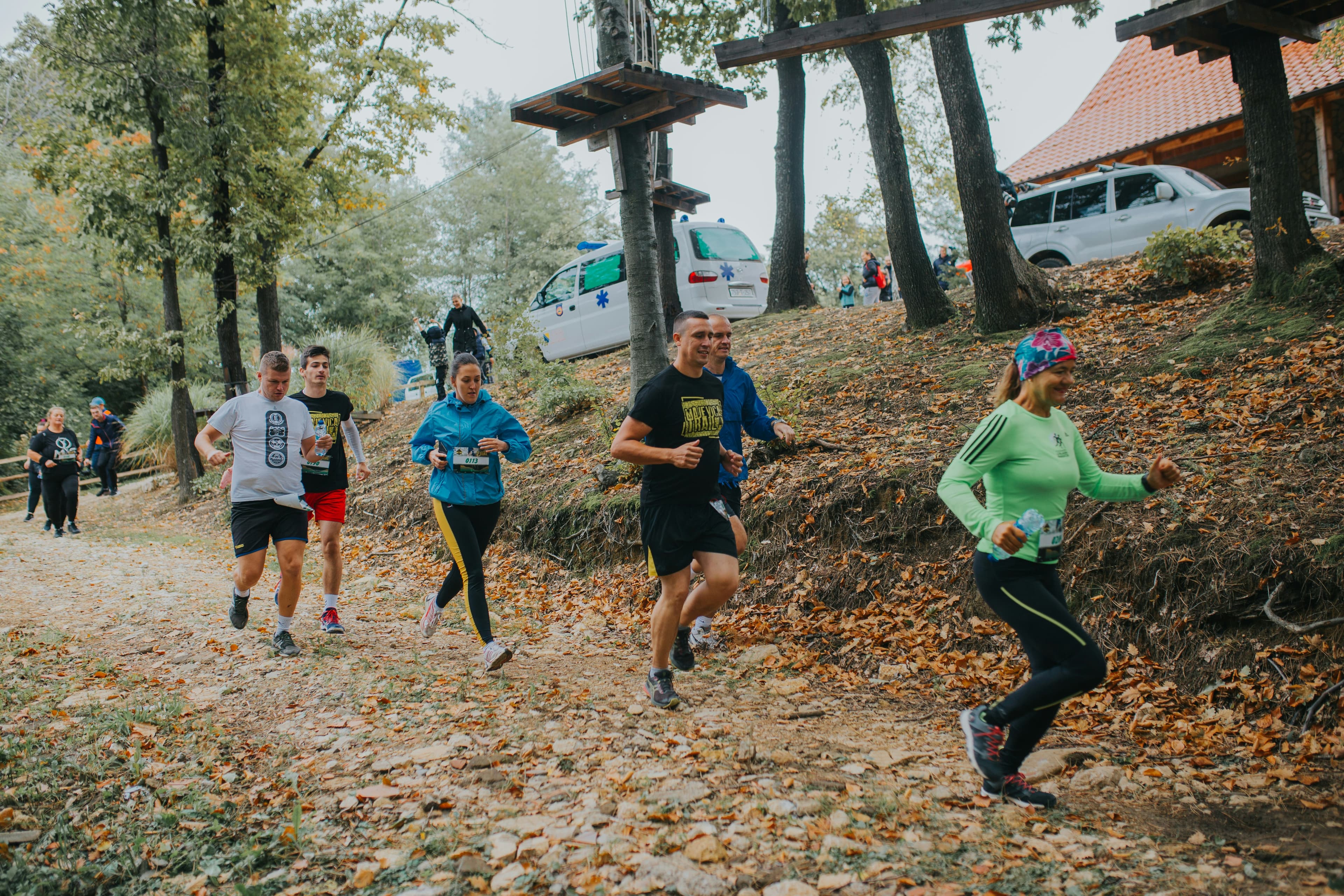 A photo of several runners running down a dirt road through the woods. The closest runner is a woman wearing a long-sleeved mint green shirt and a rainbow headband. The next runner is a man with short hair, a navy blue t-shirt, and black-and-yellow shorts. The next runner is only partially visible, but he has short hair and is wearing a navy blue pullover. The next runner is a woman in a light blue hoodie sweatshirt and black-and-yellow leggings. The next runner is a man in a light gray t-shirt, black shorts, and black sneakers. The final runner is a man in a navy blue t-shirt and red shorts.
