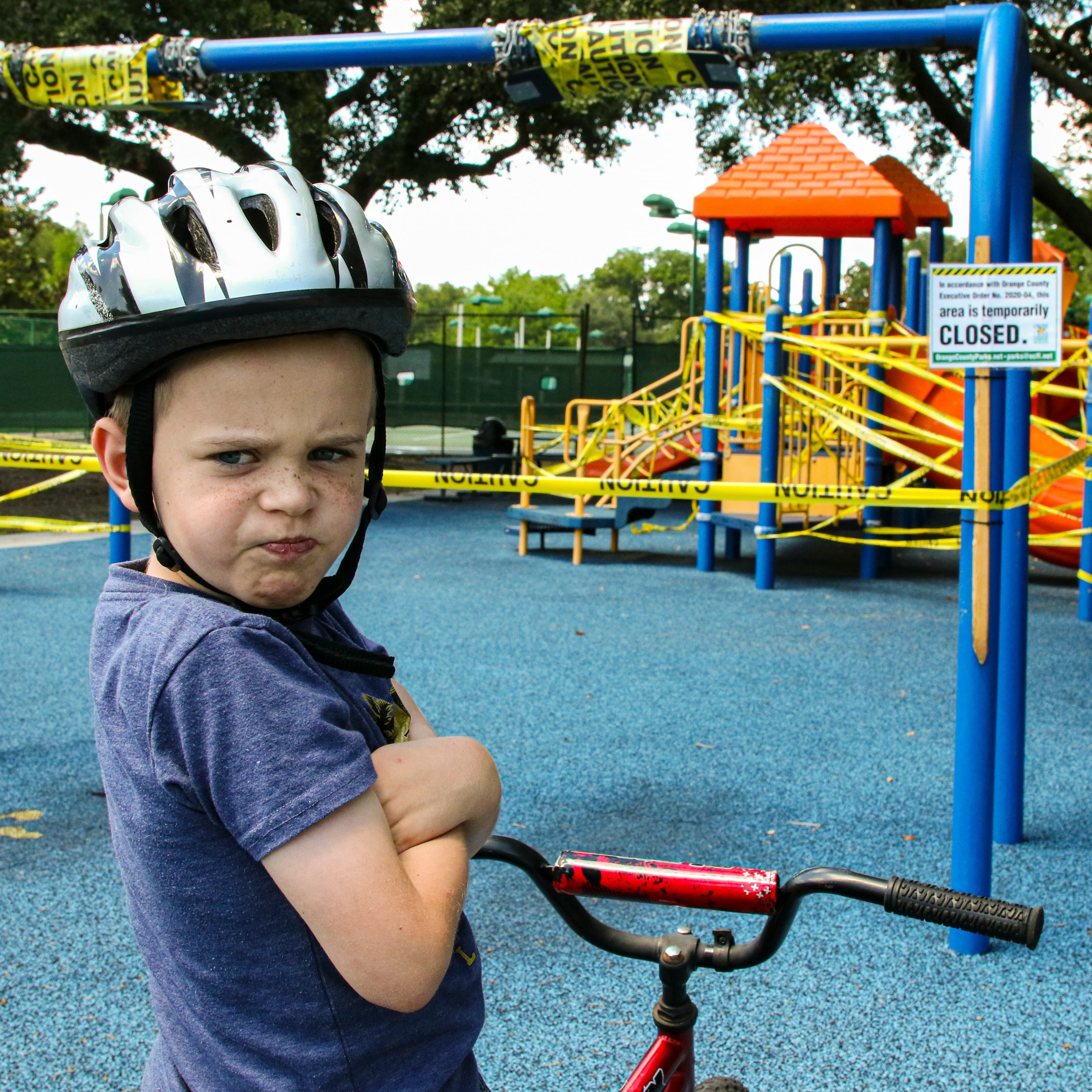 A white boy in a bike helmet stands over his bicycle and pouts with his arms crossed. In the background, we can see a playground that is wrapped in caution tape and a signs says "this area is temporarily closed."