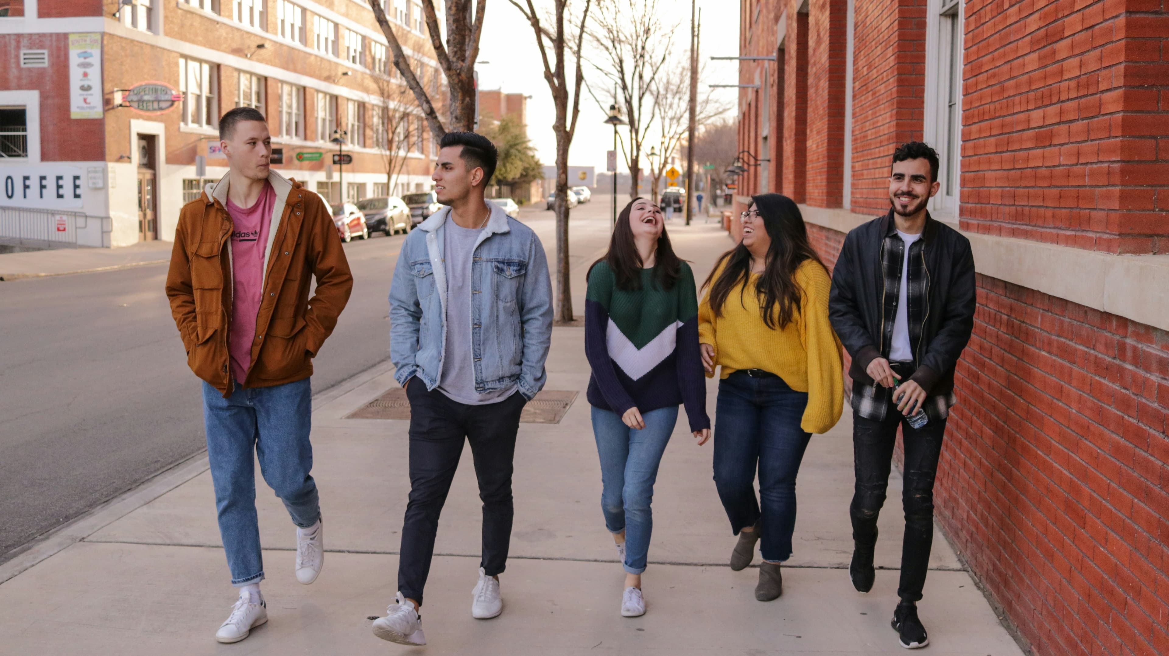 A photo of five young adults walking side-by-side down a sidewalk beside a brick building. From left to right, a man with a buzz cut in jeans, white sneakers, a soft-wash red t-shirt, and a brown coat is talking to the next man in line. He is in dark-wash jeans, white sneakers, a gray t-shirt, and a denim jacket. Next to him is a woman with long dark hair, who is throwing her head back and laughing at something the woman next to her said. She is wearing jeans, white sneakers, and a dark-colored sweater. The next woman in the line also has long dark hair, and is laughing. She is wearing a yellow sweater, dark-wash jeans, and gray shoes. Finally, at the right side of the line, a man with short curly dark hair and a goatee is smiling as well. He is wearing a dark jacket and a dark plaid shirt over a whit t-shirt, black jeans, and black sneakers and he is carrying a water bottle.