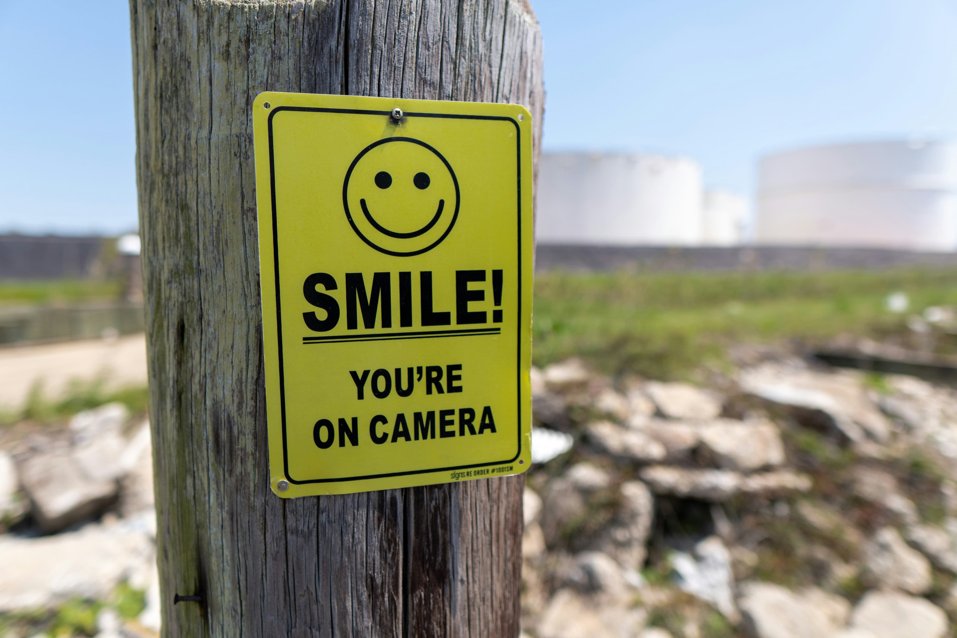 A yellow sign has a graphic of a smiley face. It reads "SMILE! You're on camera!"