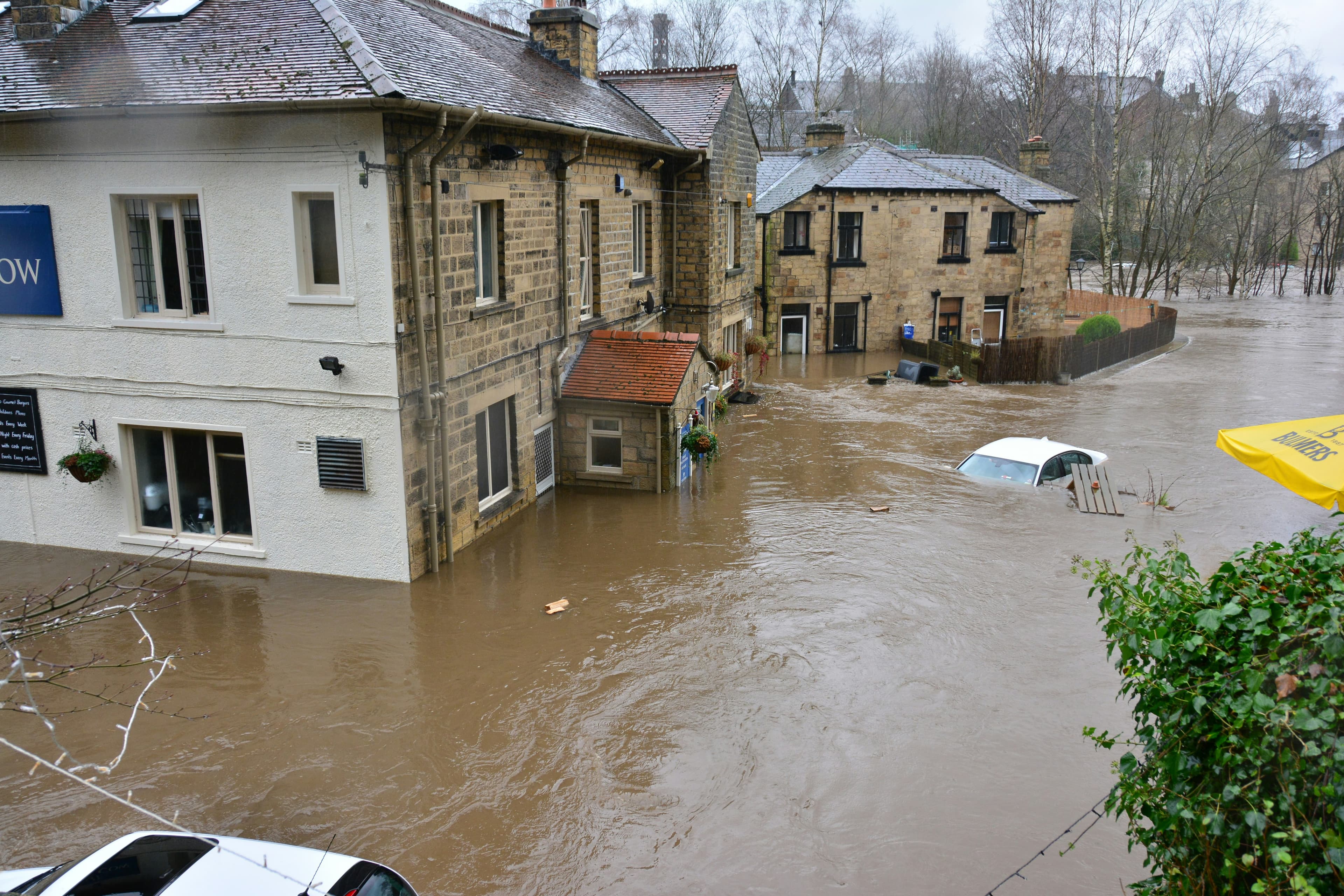 A photo of a flooded street. The water is about halfway up the first floor of the homes and has mostly covered the cars. The buildings are made of stone and have old-fashioned slate roofs.
