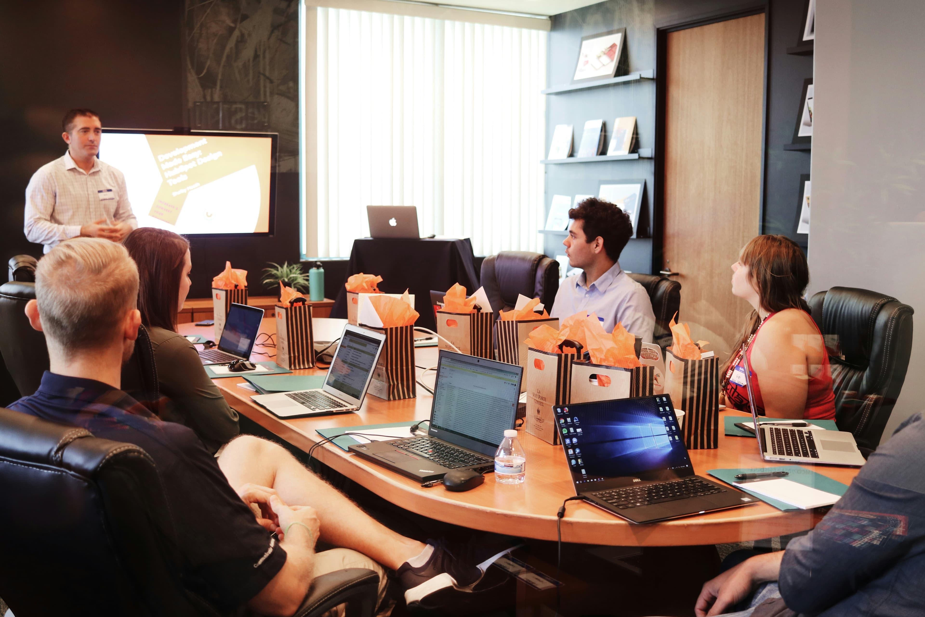 A photo of a group of people sitting around a conference table with their laptops, listening to a presentation.
