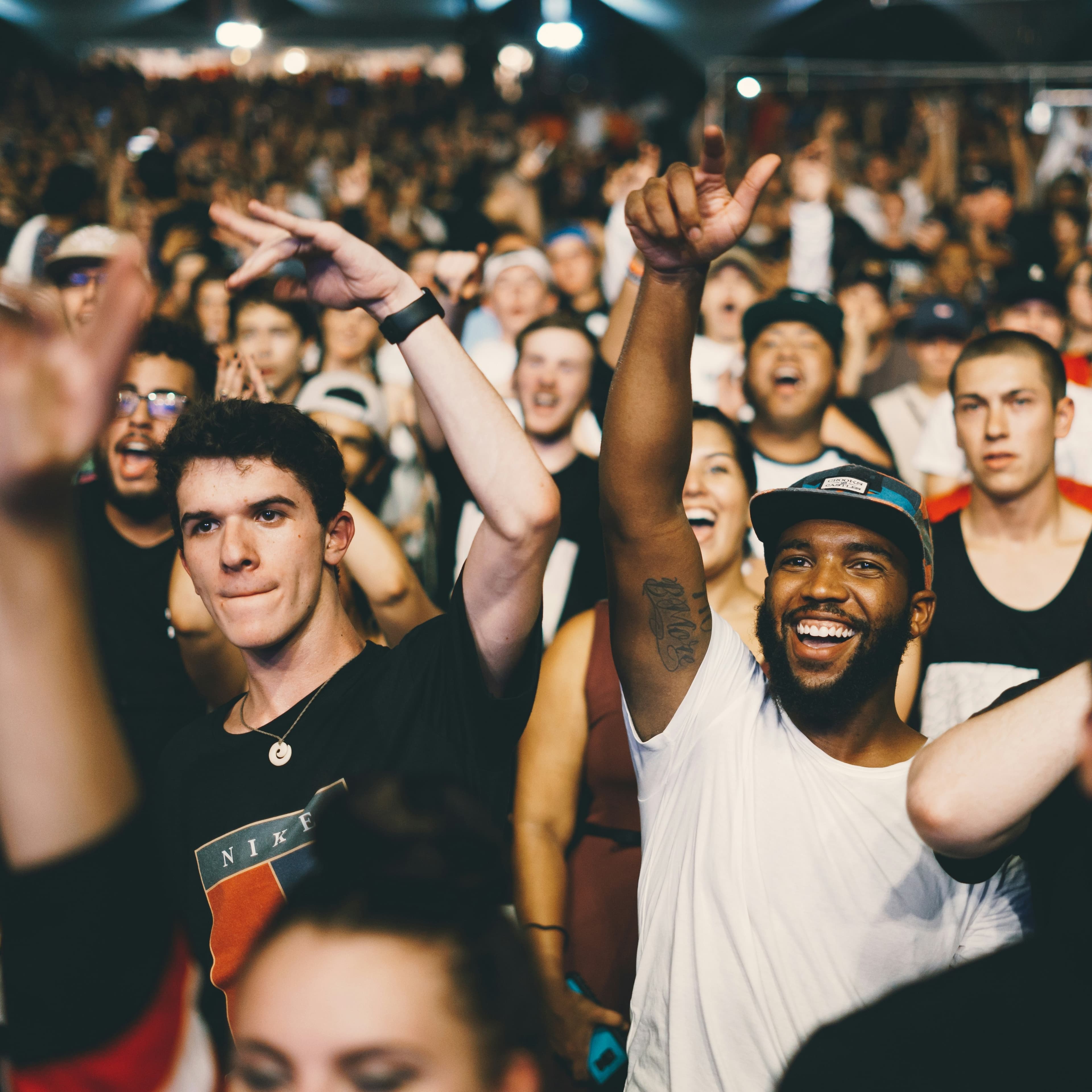 A crowd of standing people are watching a show or perhaps a sporting event. We are looking at the faces of the people and most of them look to be smiling, yelling, or otherwise looking enthusiastic. Some of them have their hands in the air and they are wearing casual clothes.