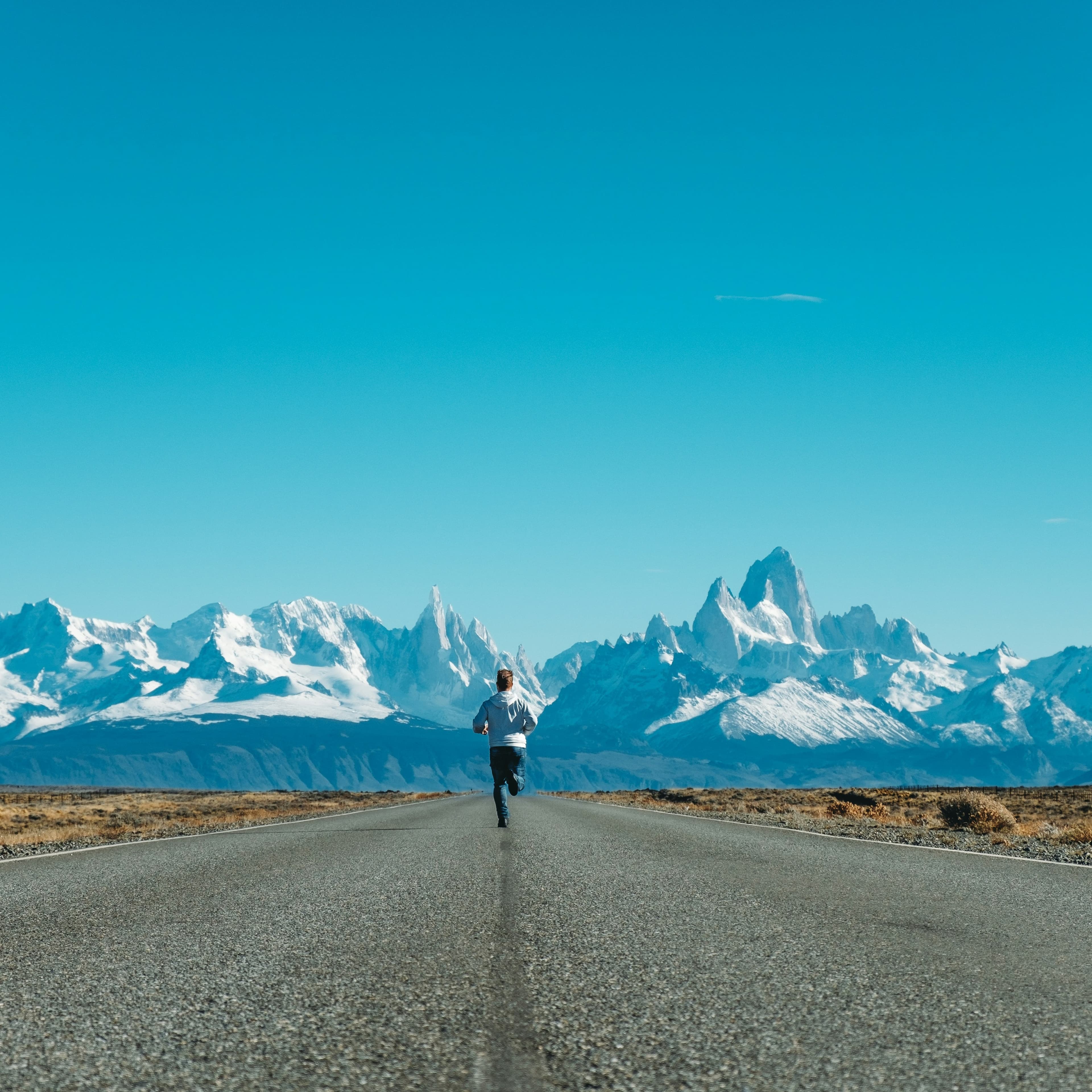 A photograph of a road stretching out in front of the camera. At the end of the road we can see a snowy mountain range. A little ways away, we can see a man running away from us, in the middle of the road.