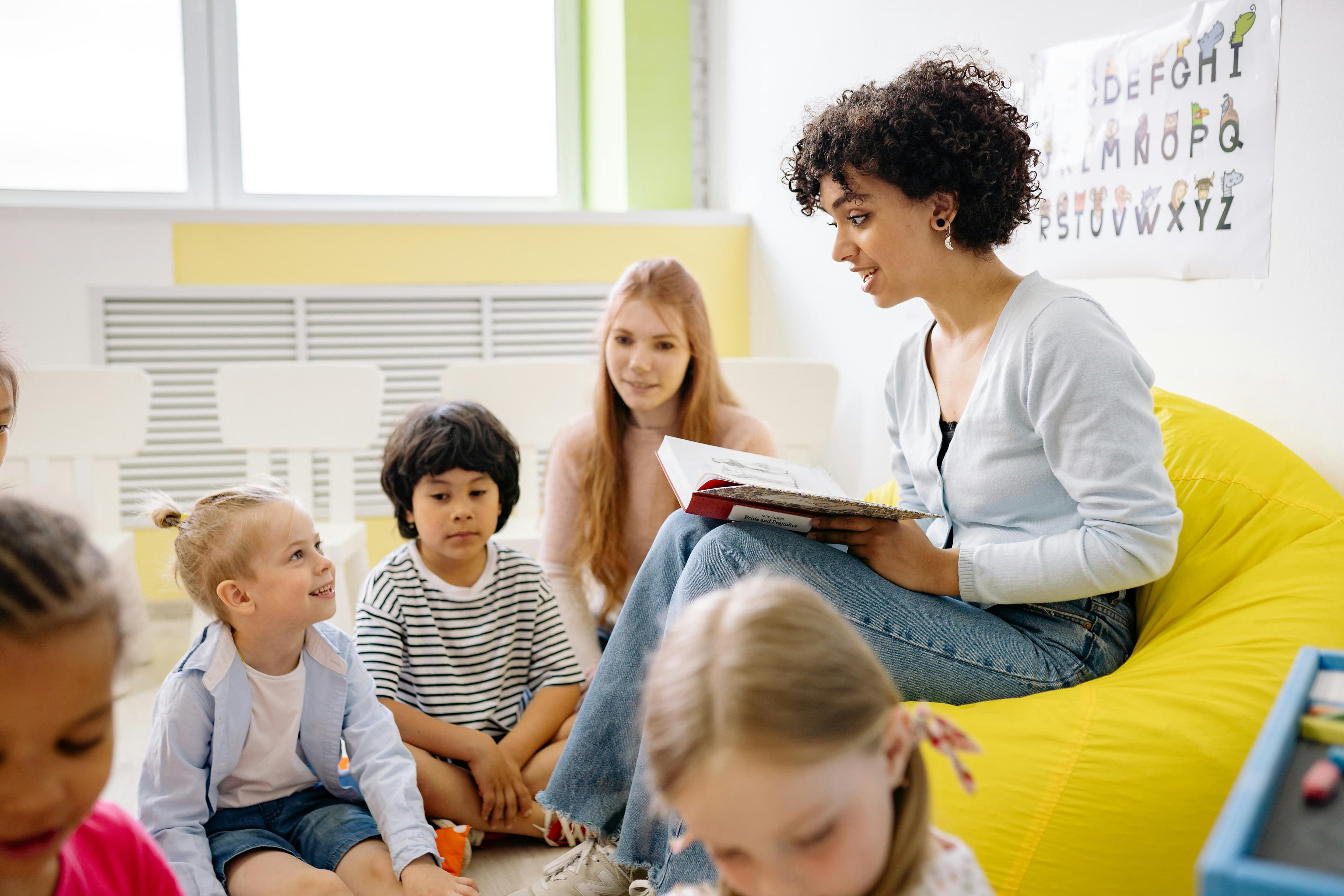A young teacher with curly hair sits on a yellow beanbag and reads a book to a classroom of young children.