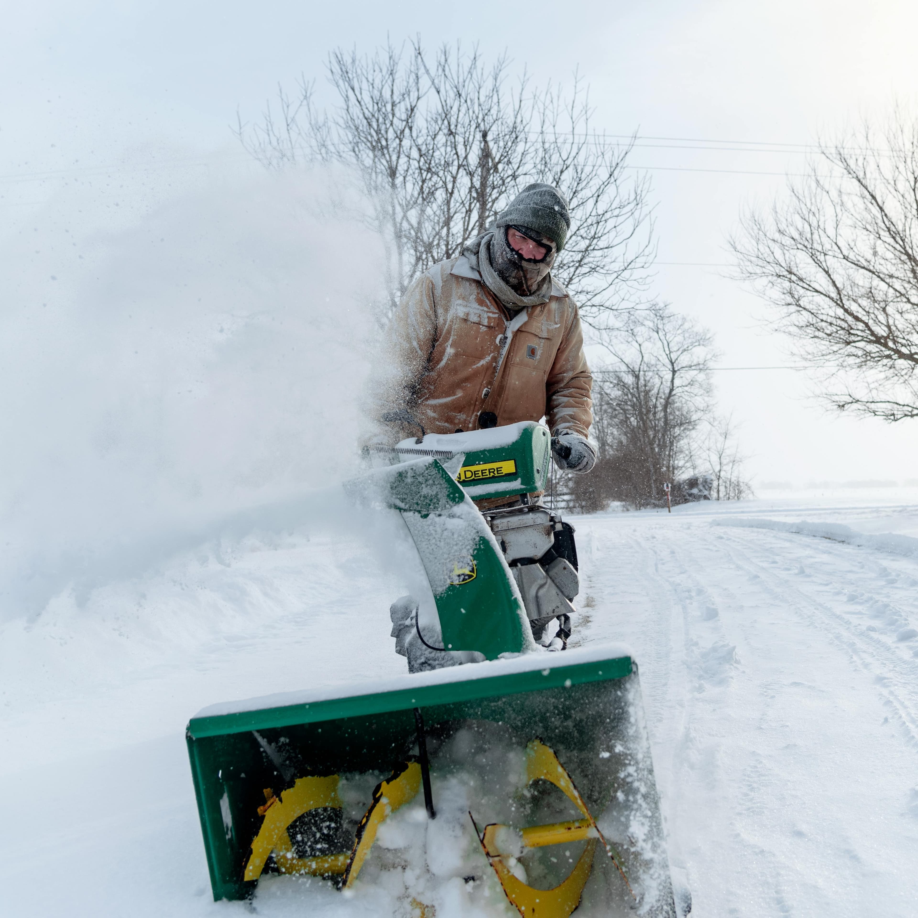 A man wearing very warm clothes is pushing a snow towards the camera. The snow is blowing off to the left of the screen.