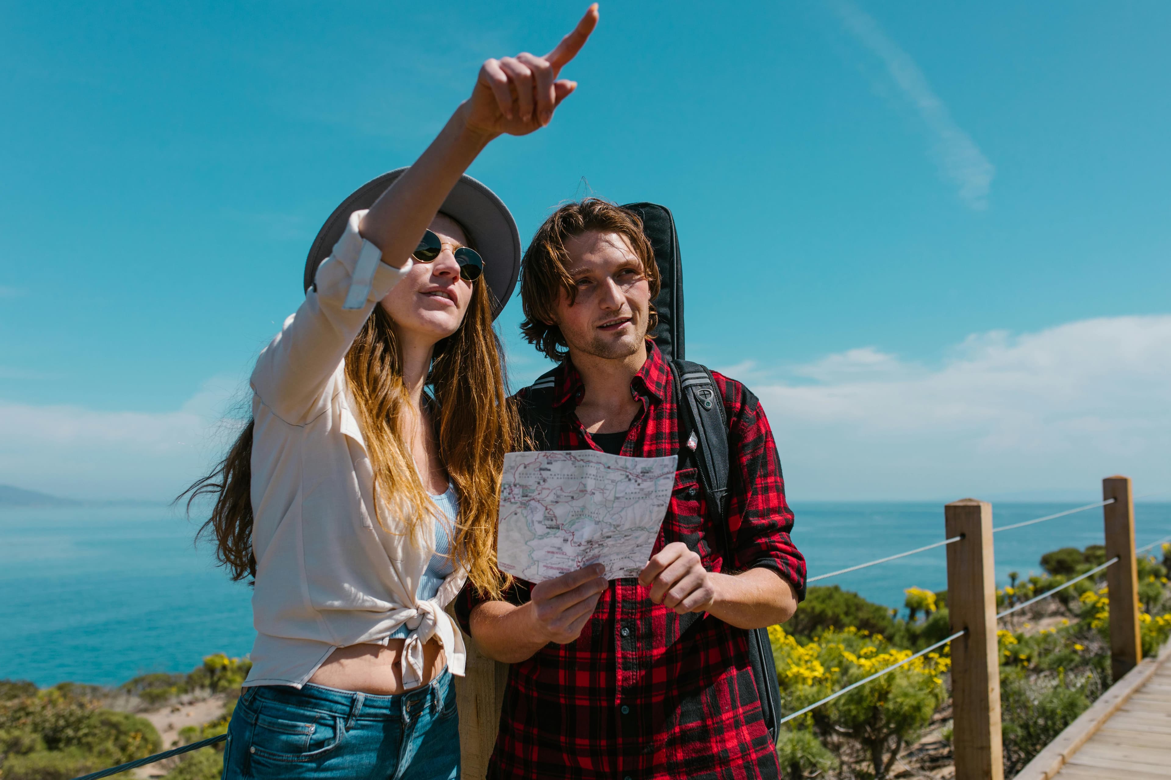 A man and woman standing on a walking path by the ocean. He is holding a map, looking at something that she is pointing to in the distance.