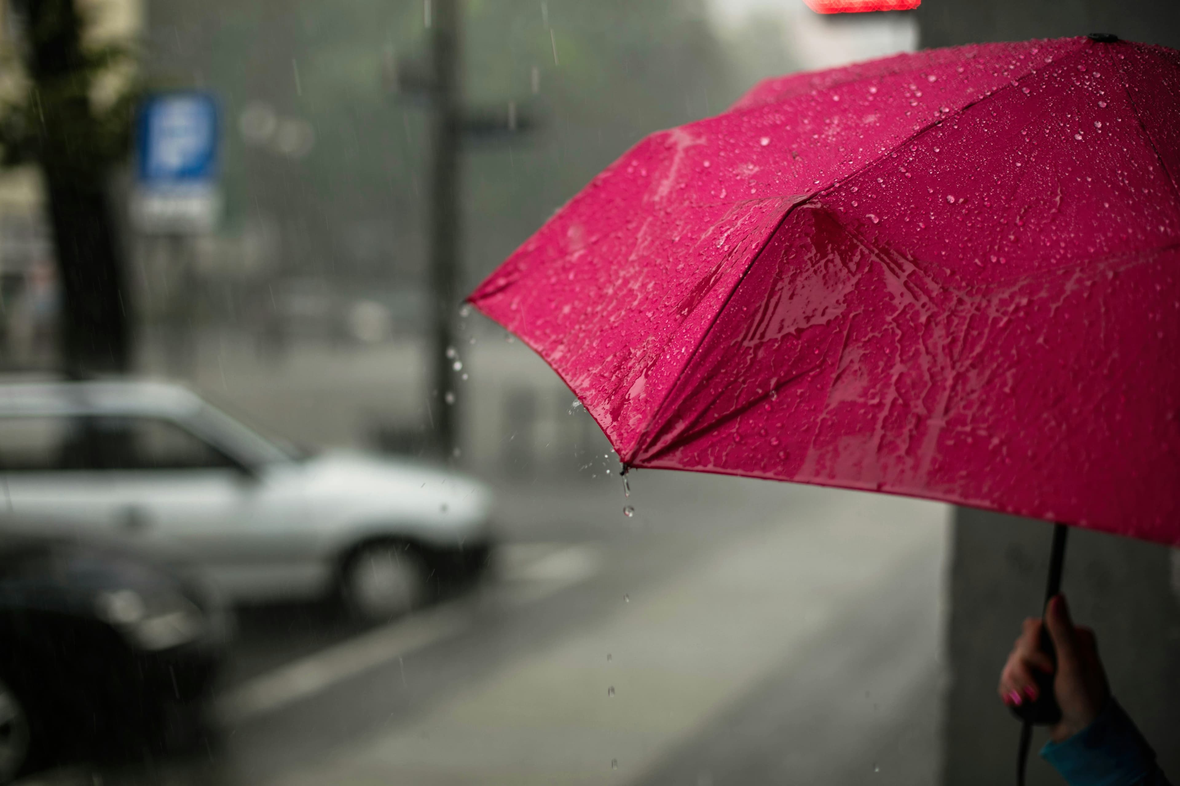 A photo of a red umbrella in the rain. We can see a woman's hand holding the umbrella but the person holding it is out of frame.