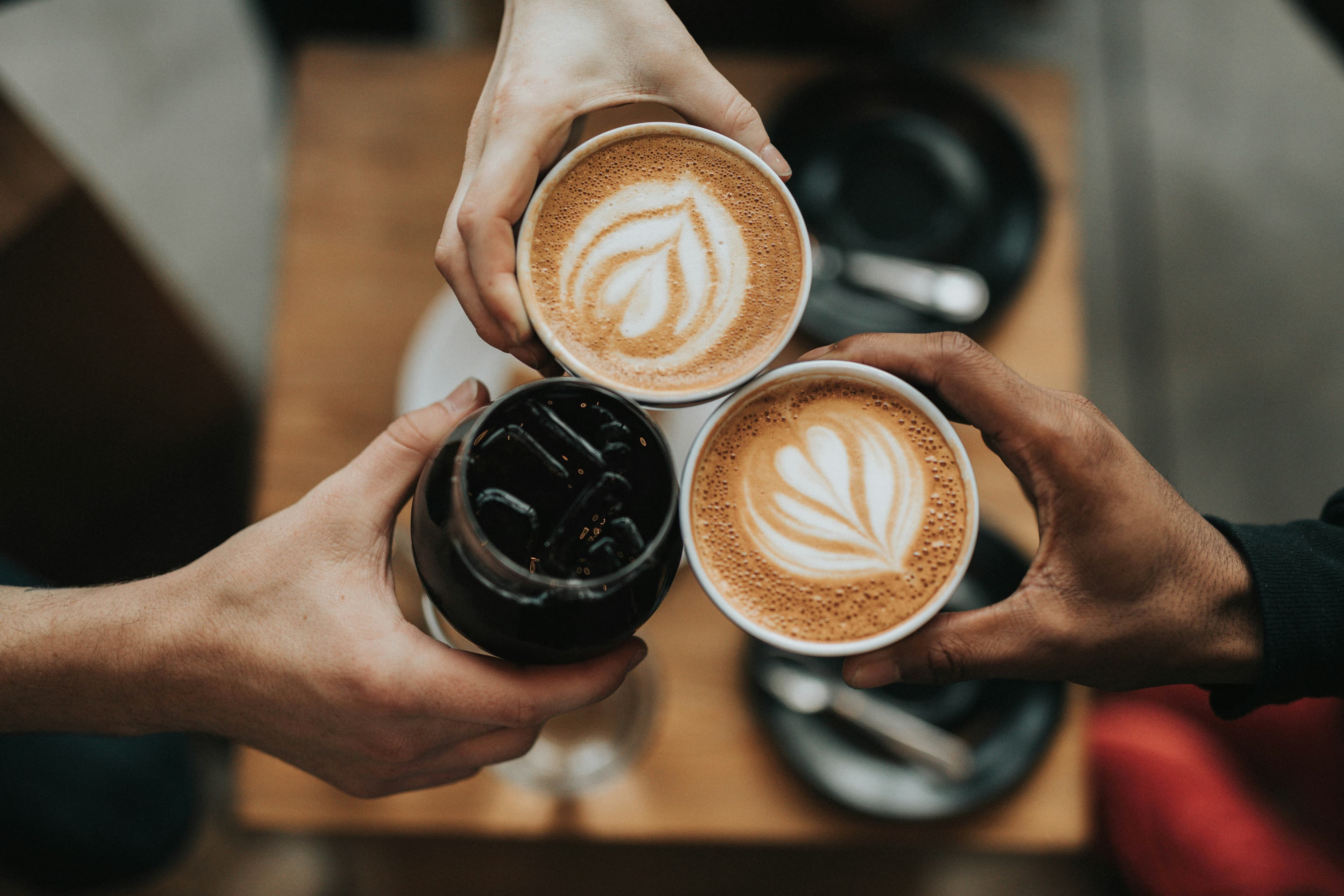 A photo of three cups of coffee taken from above. Three different hands are holding the cups and they meet in the middle. Two coffees are cappuccinos with designs in the cream and one is an iced black coffee.