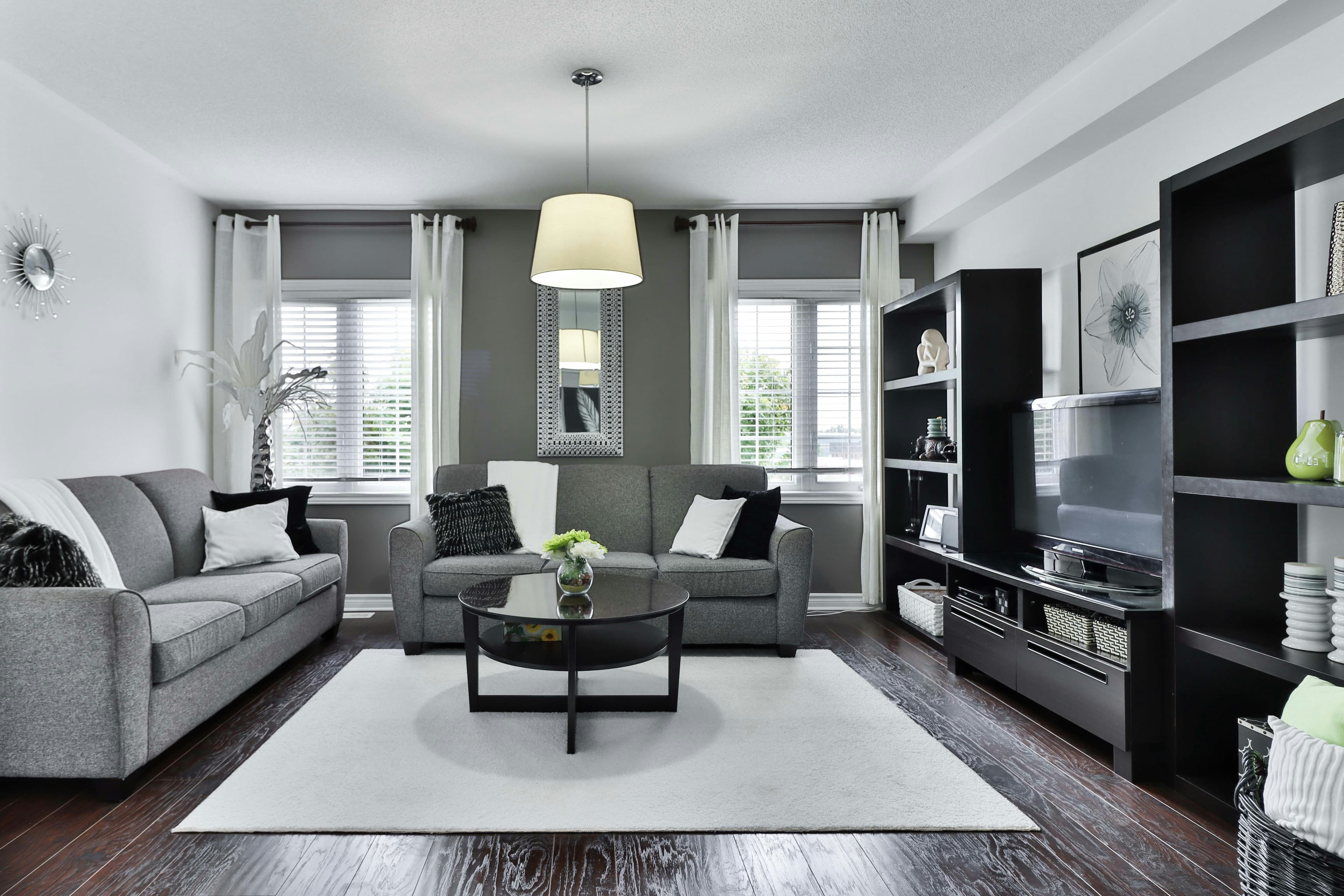 A photo of a modern living room decorated in gray, black, and white. There are two couches, positioned on the far side and the left side of a square right rug. In the middle of the rug is a round black coffee table with a small vase on it. A hanging lamp is centered over the coffee table. On the wall behind the couch on the opposite side of the room are two windows with white curtains. Positioned between the two windows is a tall mirror with a white frame. Both couches have black and white cushions, and a white blanket is draped on the back of the sofa on the left. Above this sofa a silver and white clock hangs on the wall. On the right side of the room are two black book cases and, between them, a television sitting on a black console. The book cases contain decorative objects. A black and white painting of a flower hangs over the TV. We can just see the corner of a bin of throw pillows positioned just to the right of the camera.